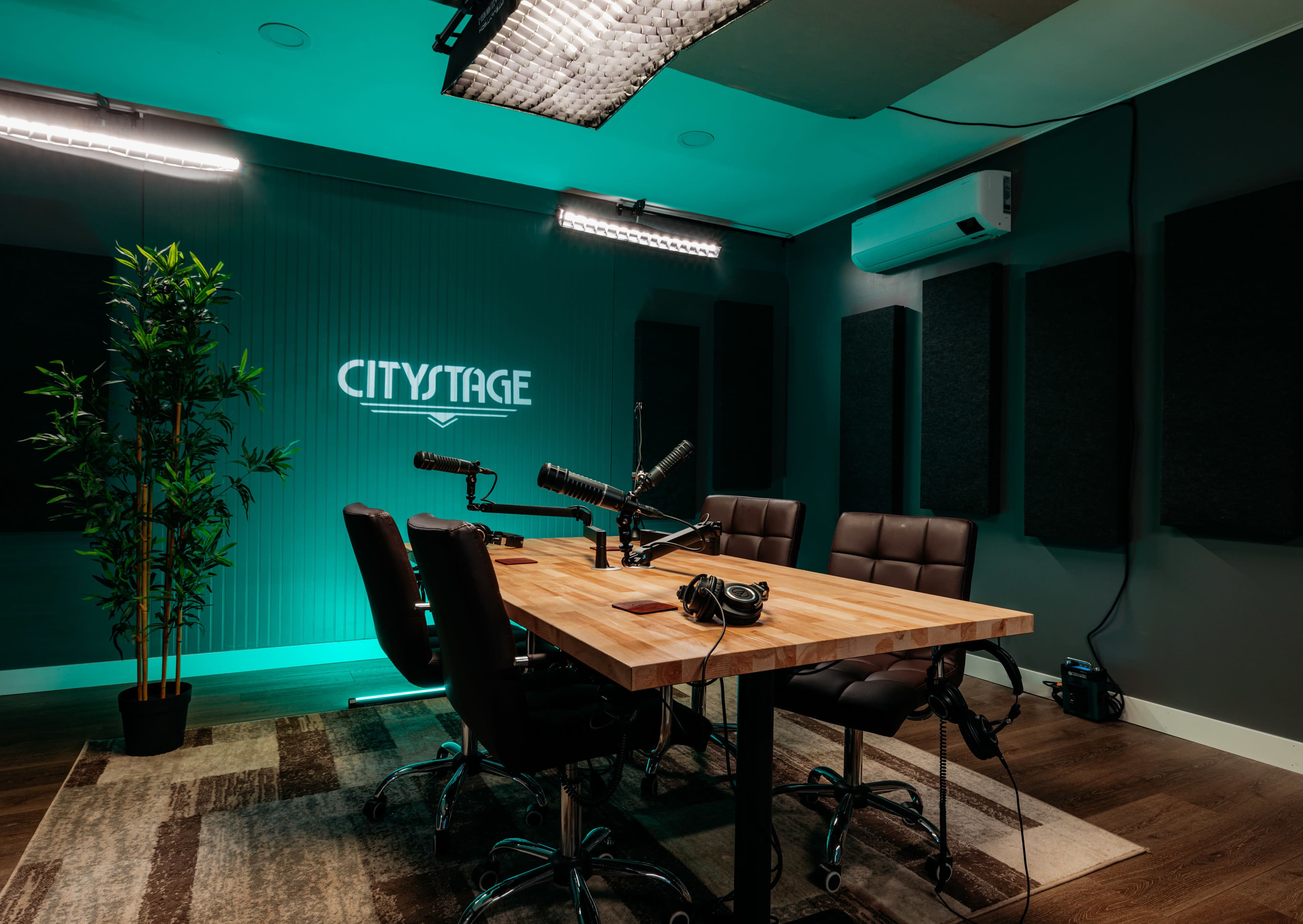 A podcast recording studio features a wooden table surrounded by leather chairs, with microphones and headphones set up, and soundproof panels on the walls.