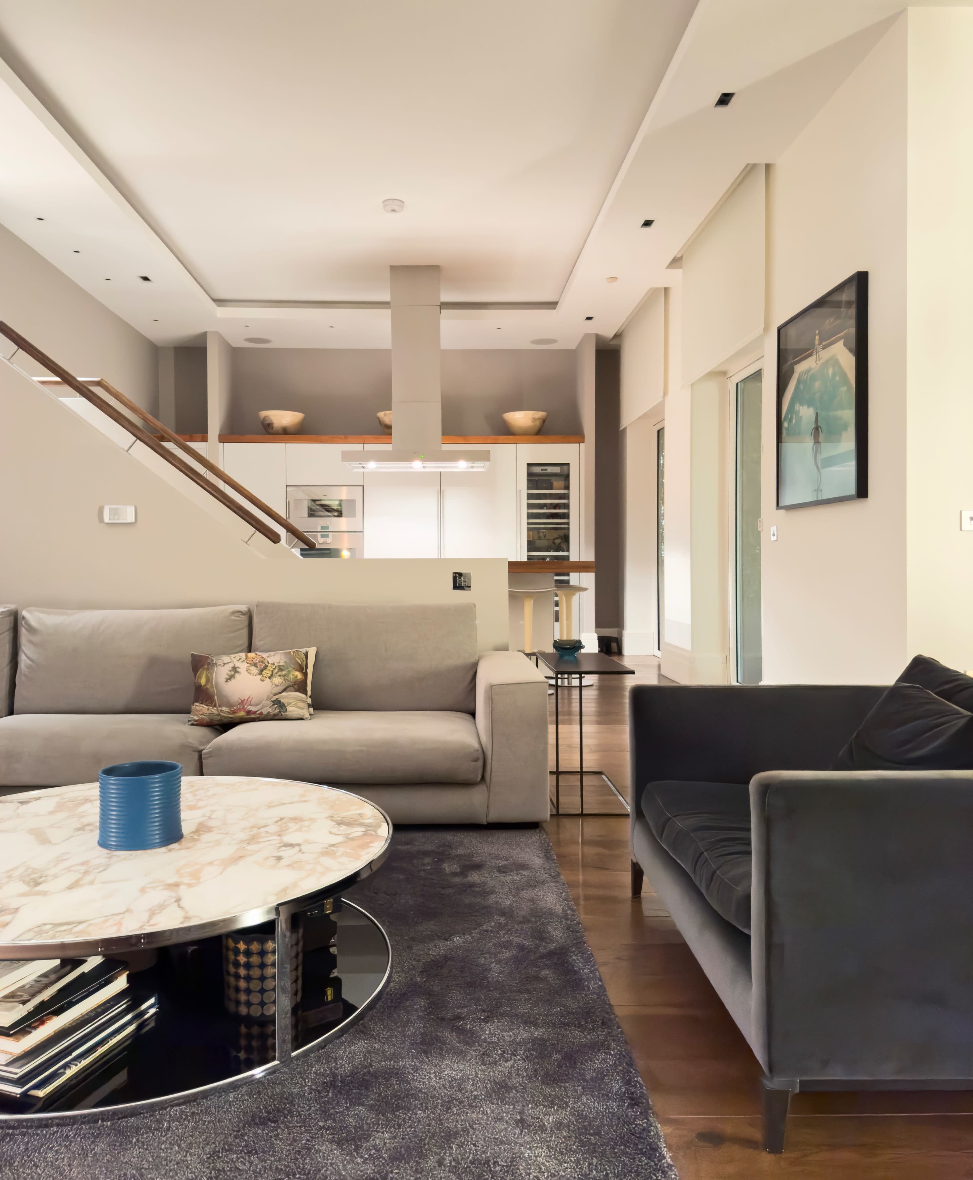 The image shows a modern living room featuring a gray sofa, a darker armchair, and a round marble coffee table set on a soft rug, with a kitchen area visible in the background.