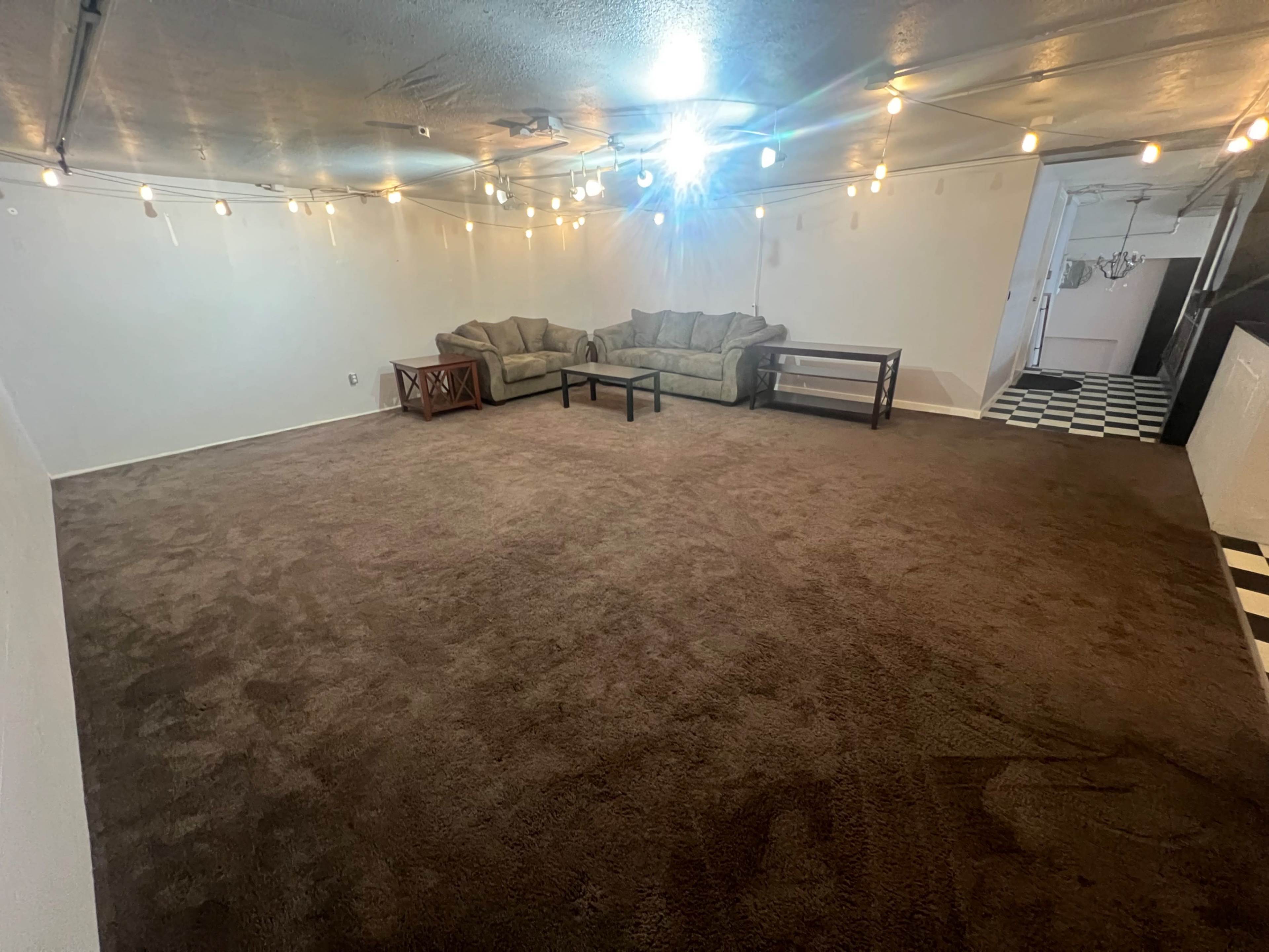 A spacious room with carpeted flooring, featuring a seating area with two couches, a coffee table, and string lights overhead.