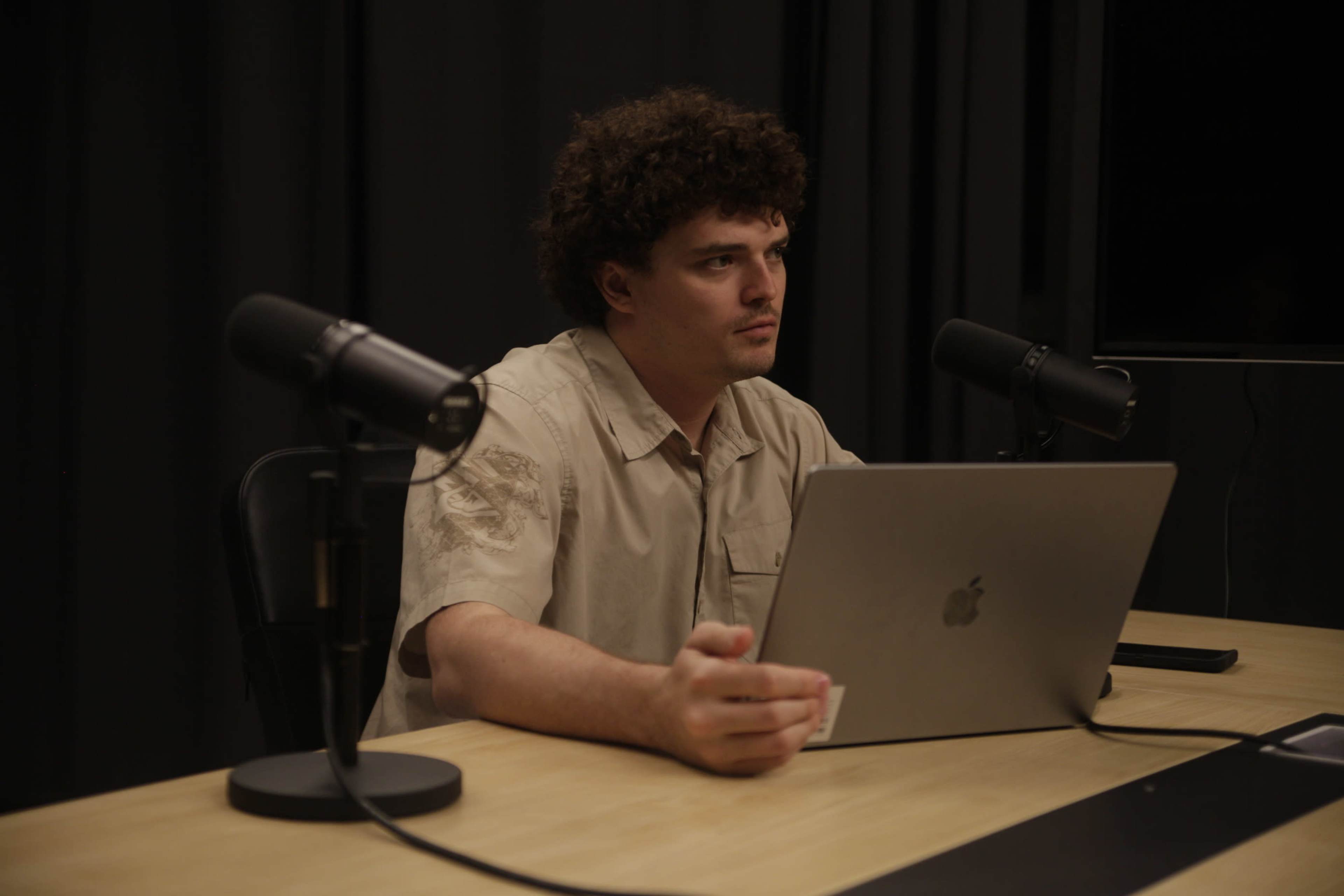 A man with curly hair sits at a table with a laptop and two microphones in front of him, against a backdrop of black curtains.