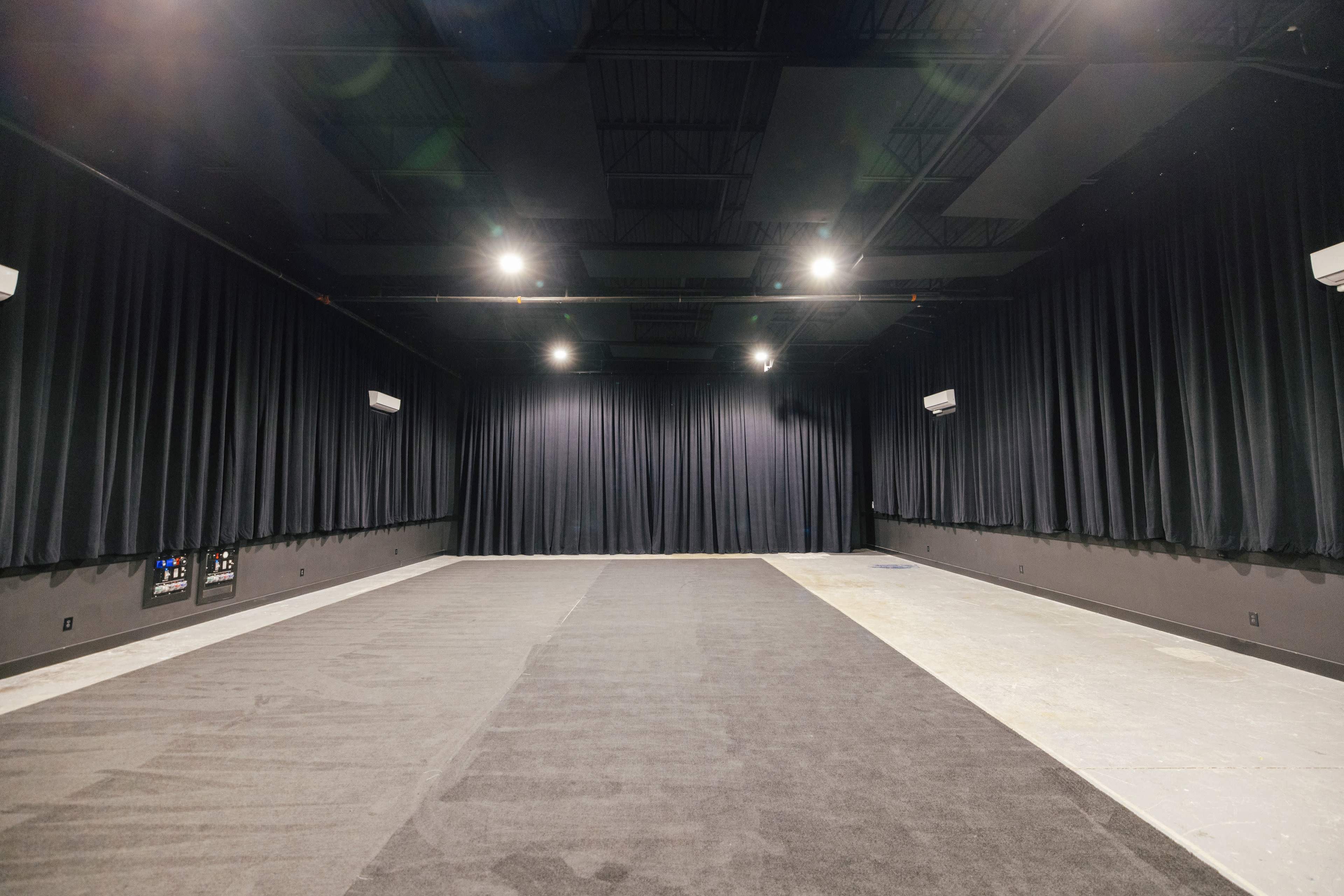 The image shows a large, empty room with dark curtains on the walls and several overhead lights illuminating the space.