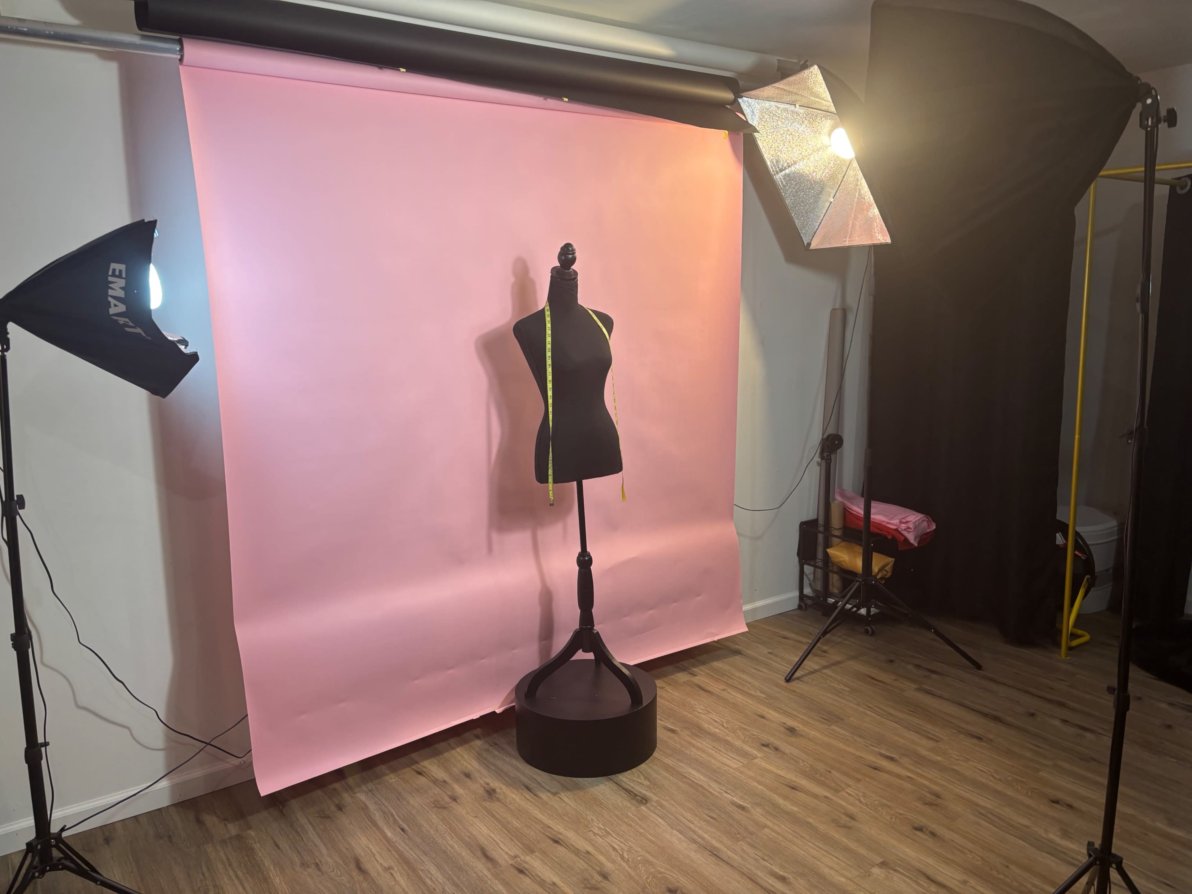 The image shows a photography studio setup with a pink backdrop, a dress form on a stand, and lighting equipment arranged around it.