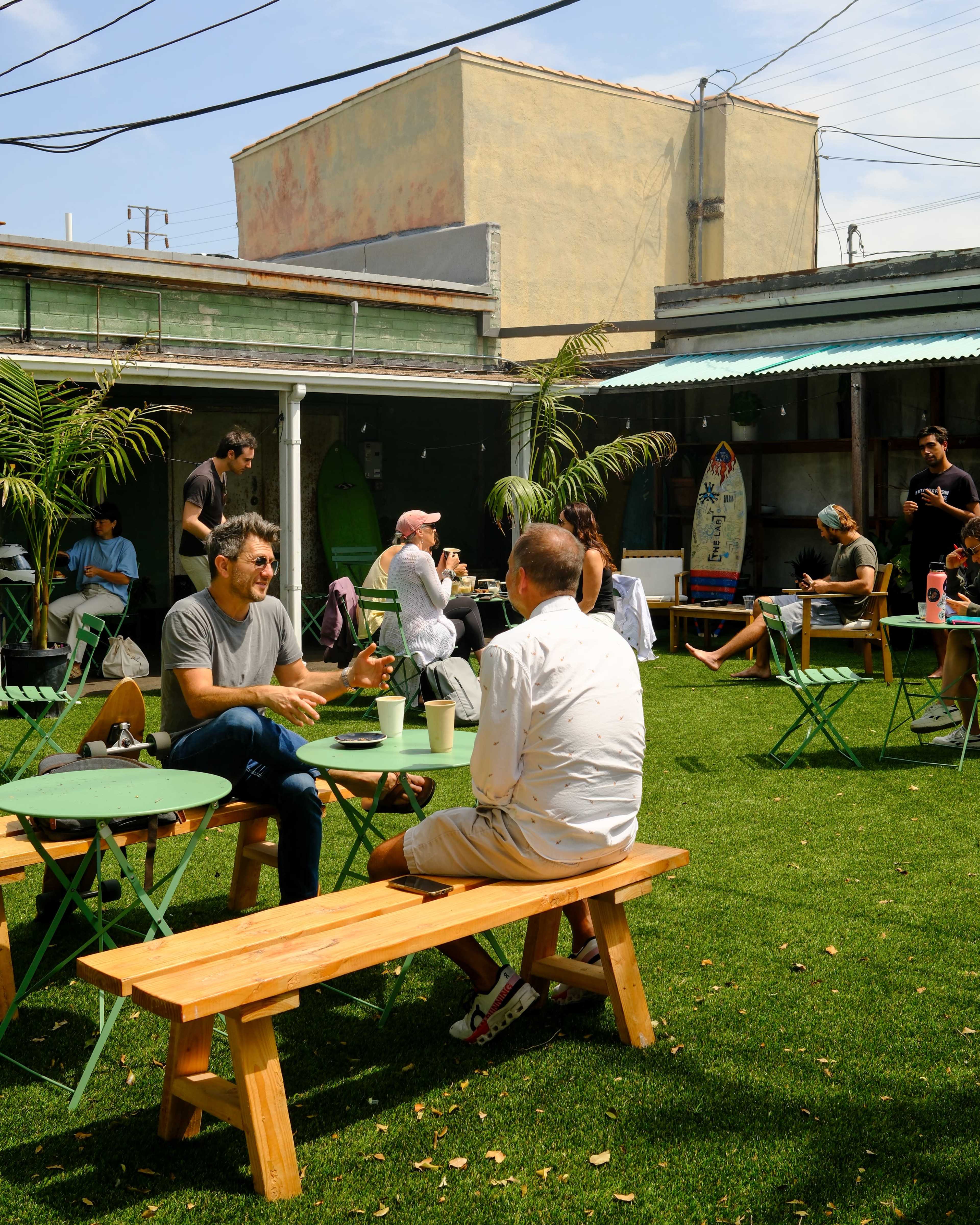 Two men sit at a wooden table in a green courtyard area filled with picnic tables, surrounded by people enjoying their time in the sun.