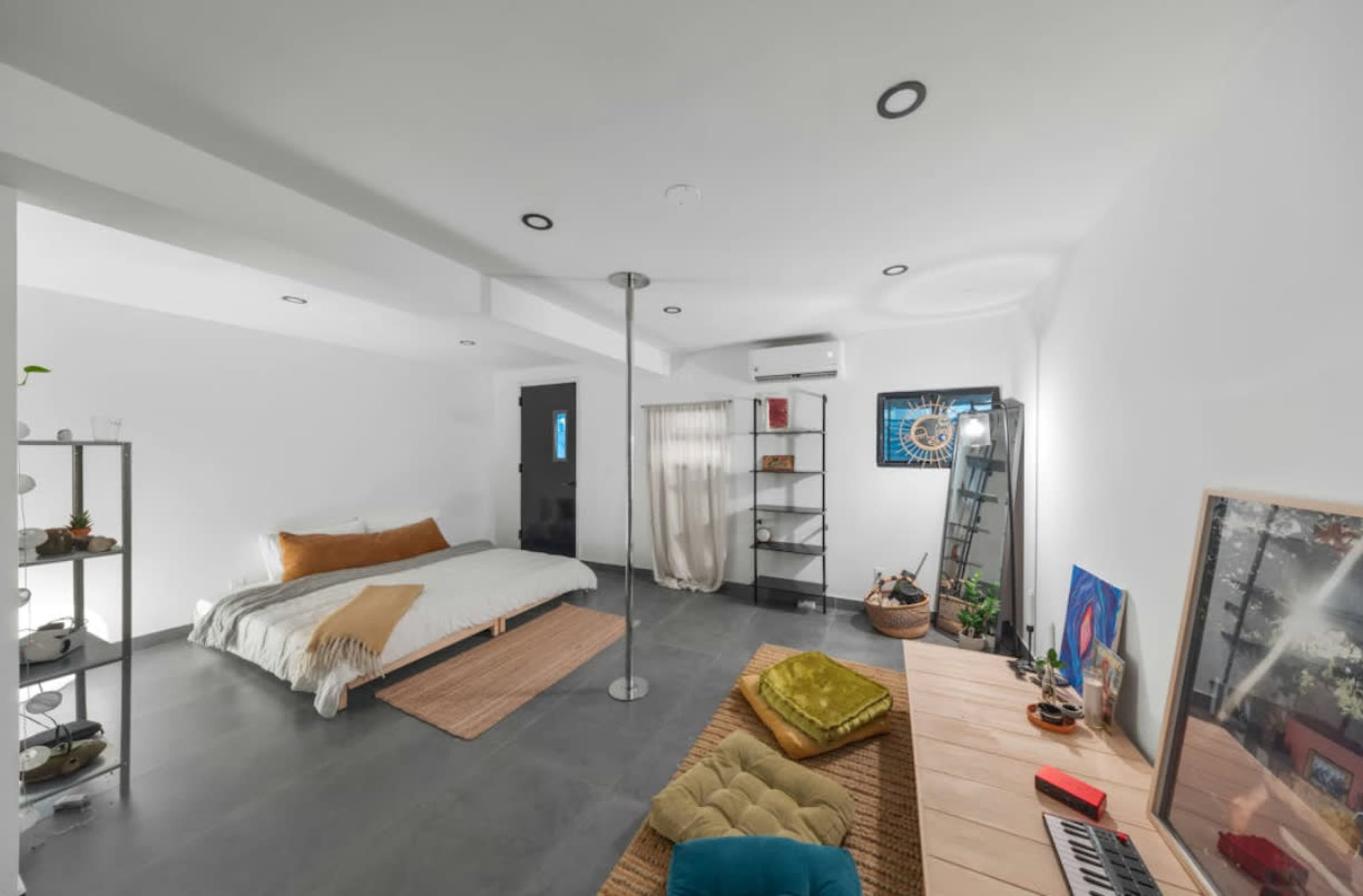 The image displays a minimalistic bedroom featuring a bed, a shelf with plants, and a wooden table with a keyboard, set in a spacious, well-lit room.