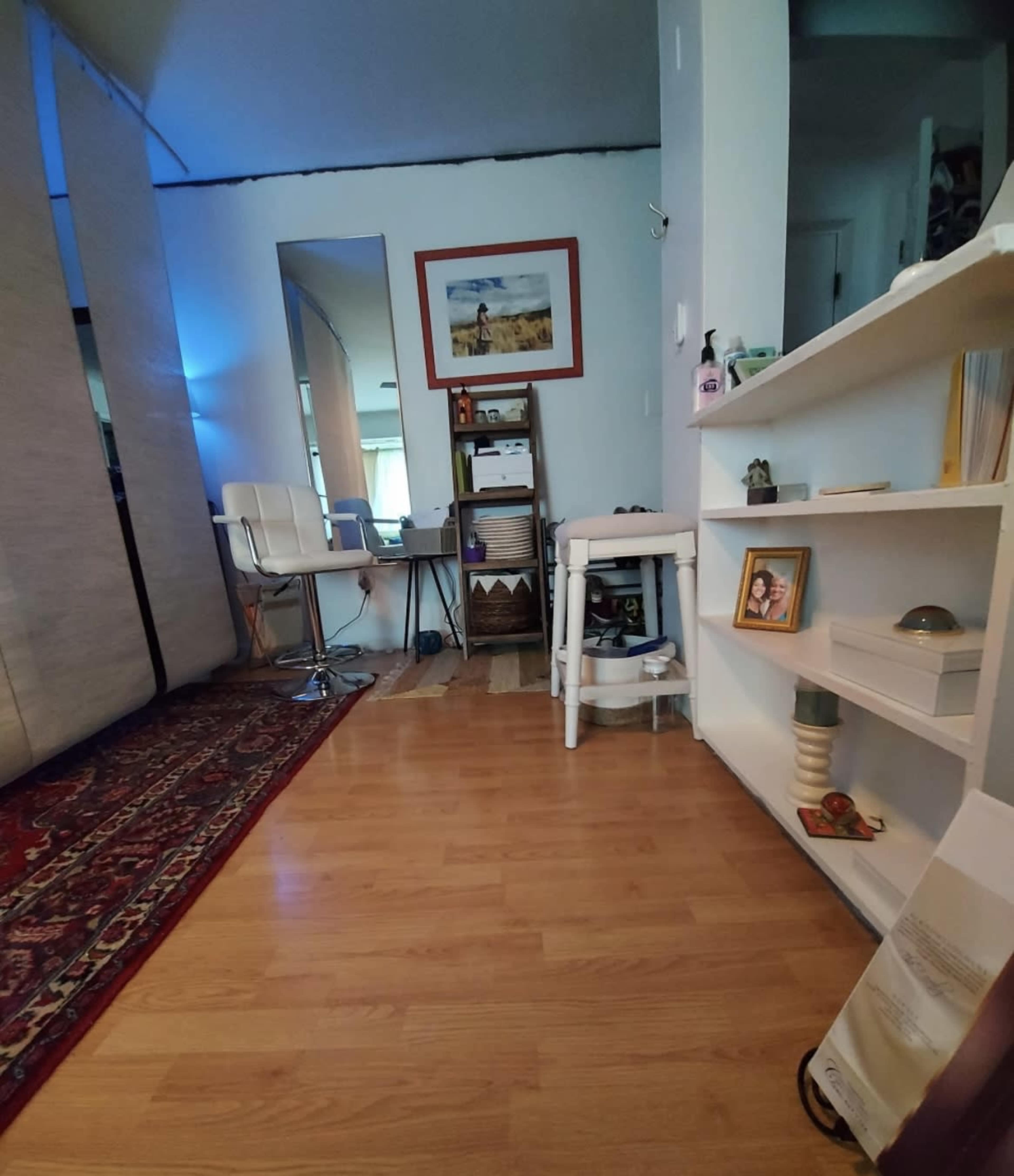 A small, wooden-floored room features a desk with a chair, a framed picture on the wall, and a bookshelf filled with various items.
