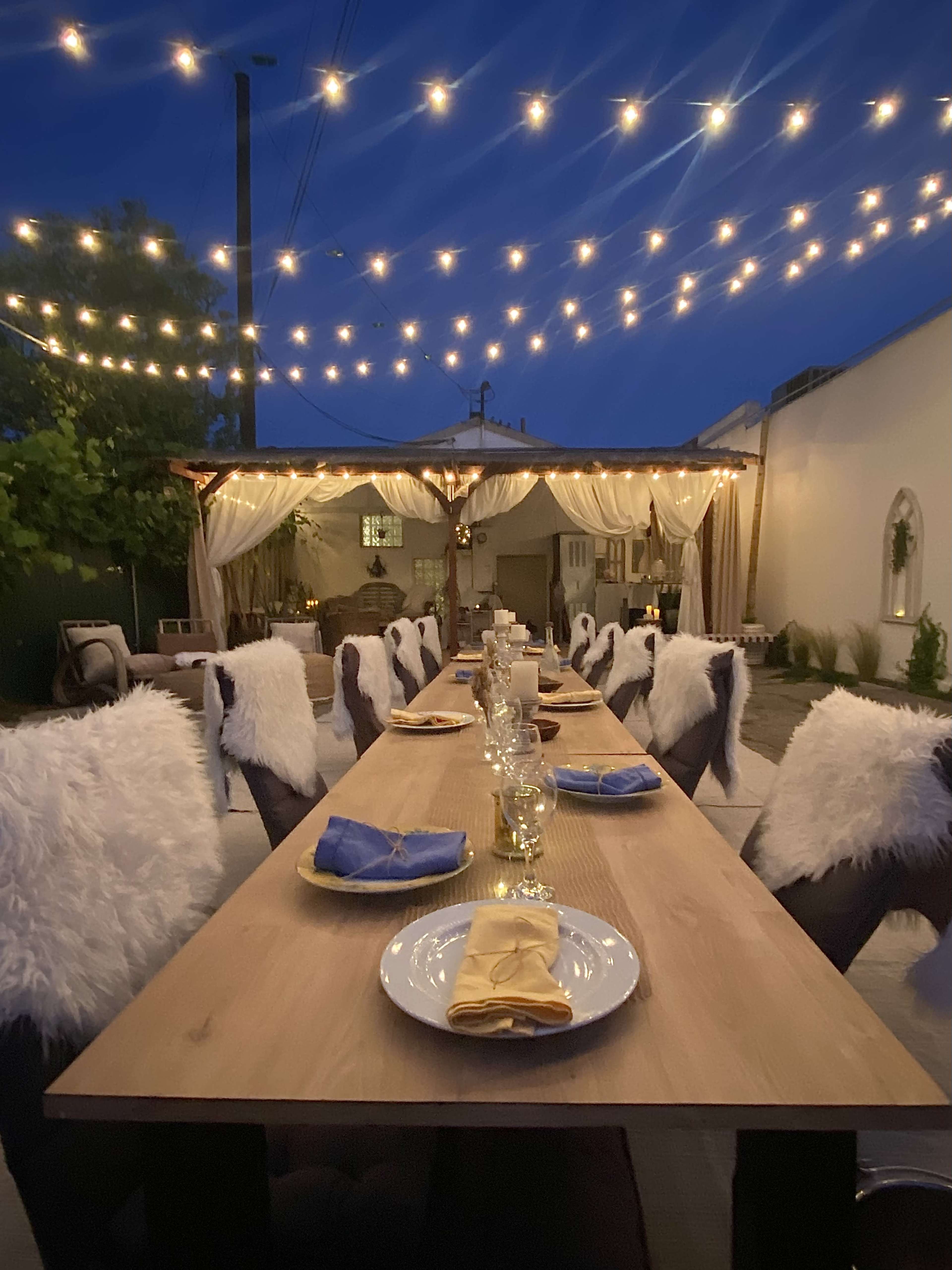 A long wooden dining table is set with plates and utensils under string lights in an outdoor setting at night.