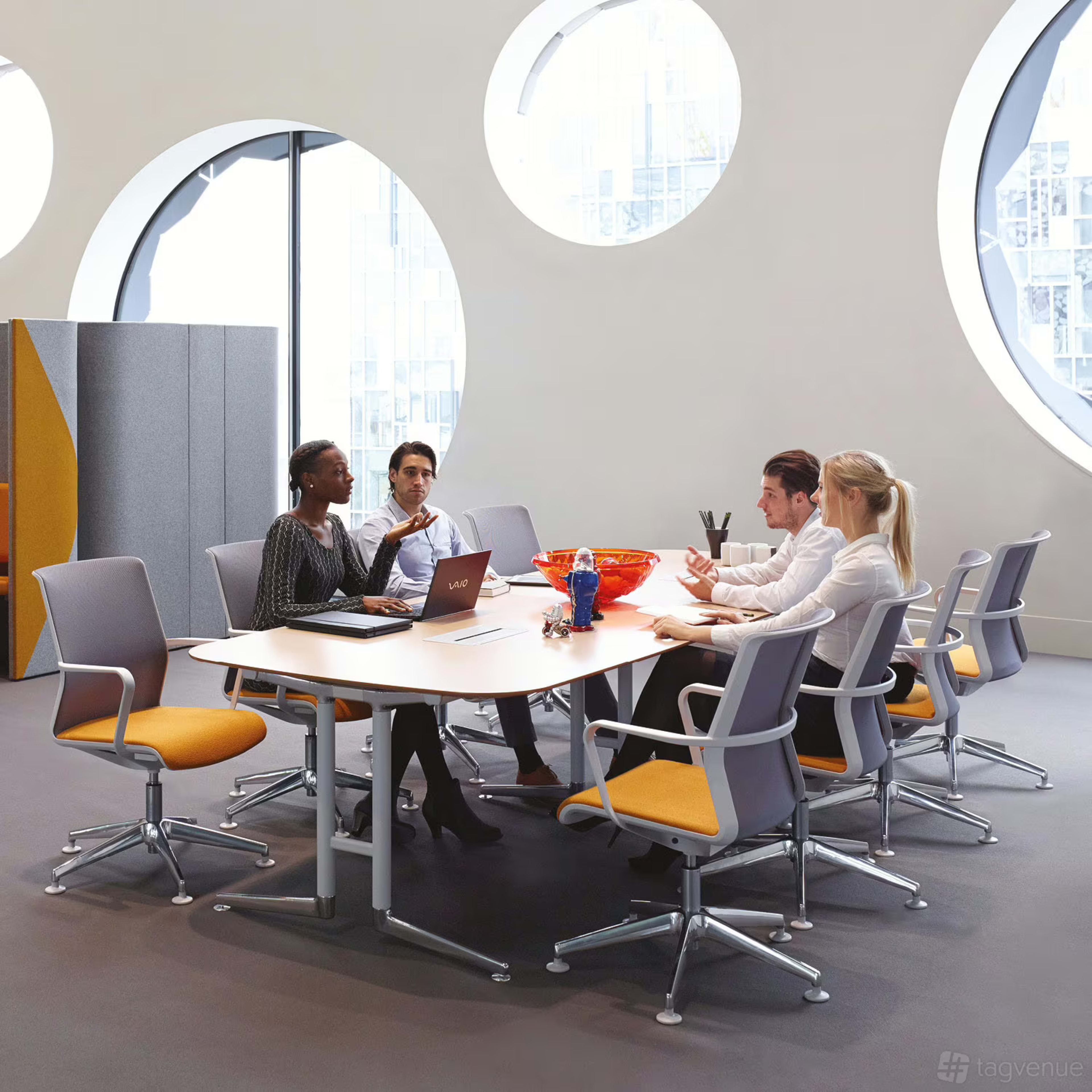 A group of four individuals is seated at a conference table in a modern office space with large circular windows.