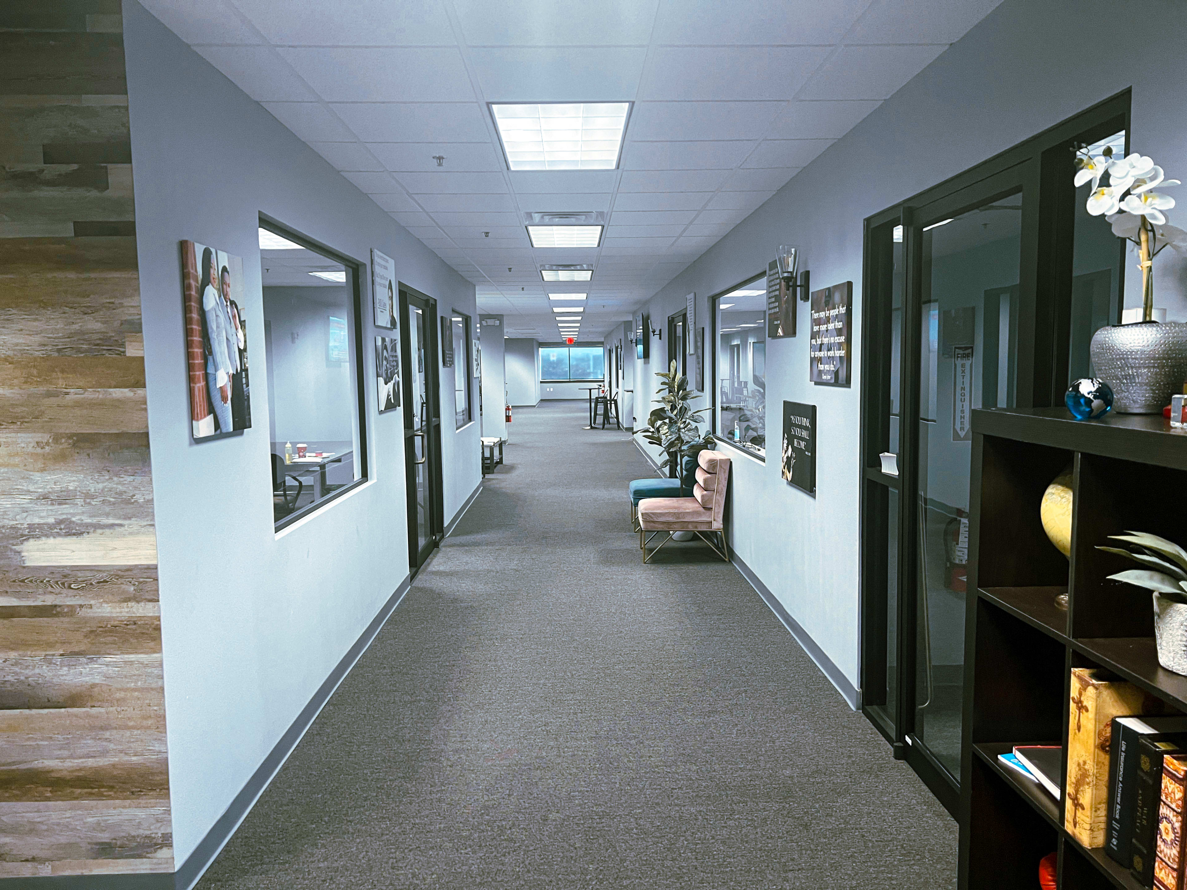 The image shows a long hallway in an office building with several doors on either side and seating areas along the walls.