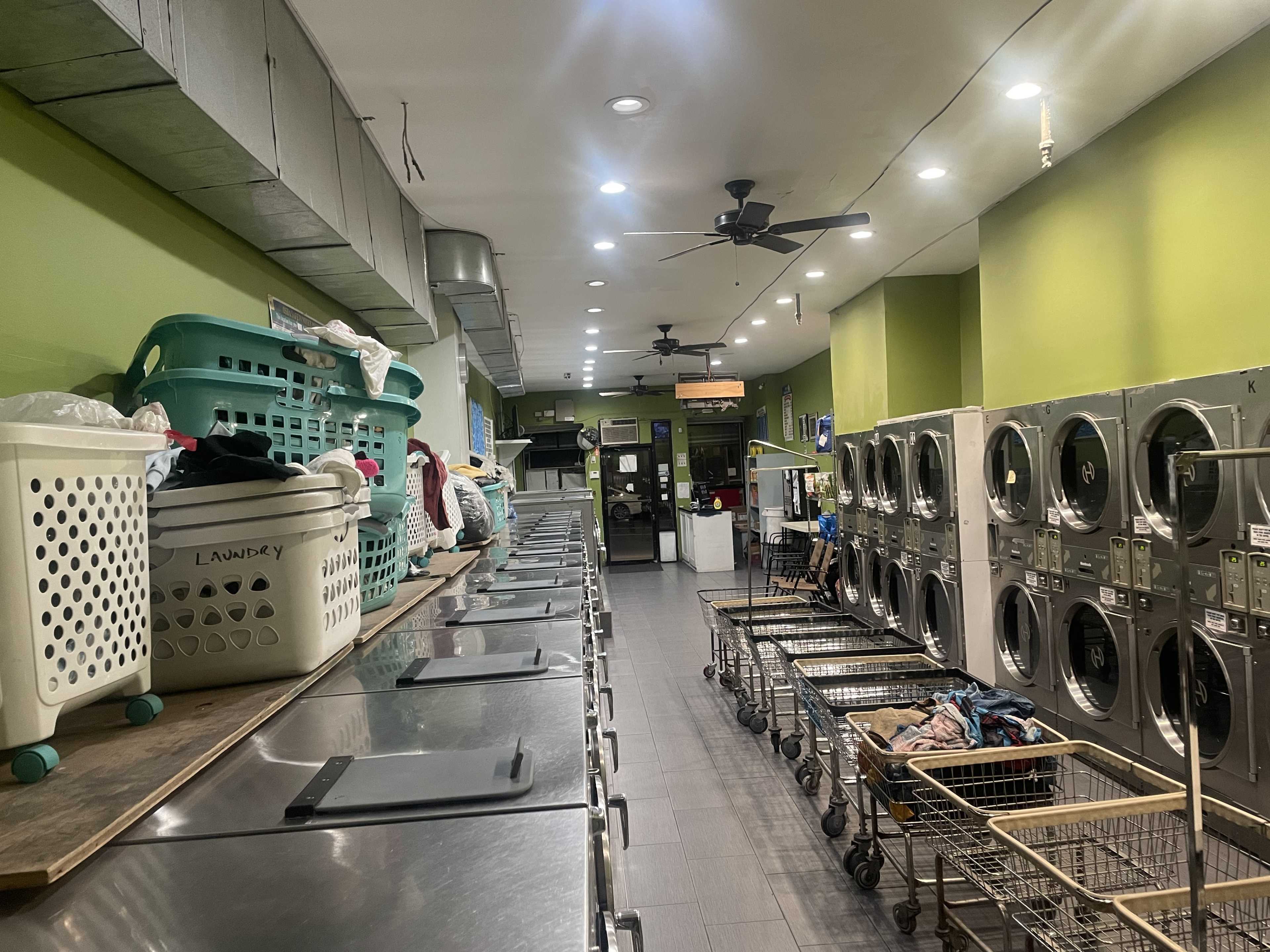 A laundromat with washing machines and dryers lined up on one side, while laundry baskets and carts are arranged along a counter on the opposite side.