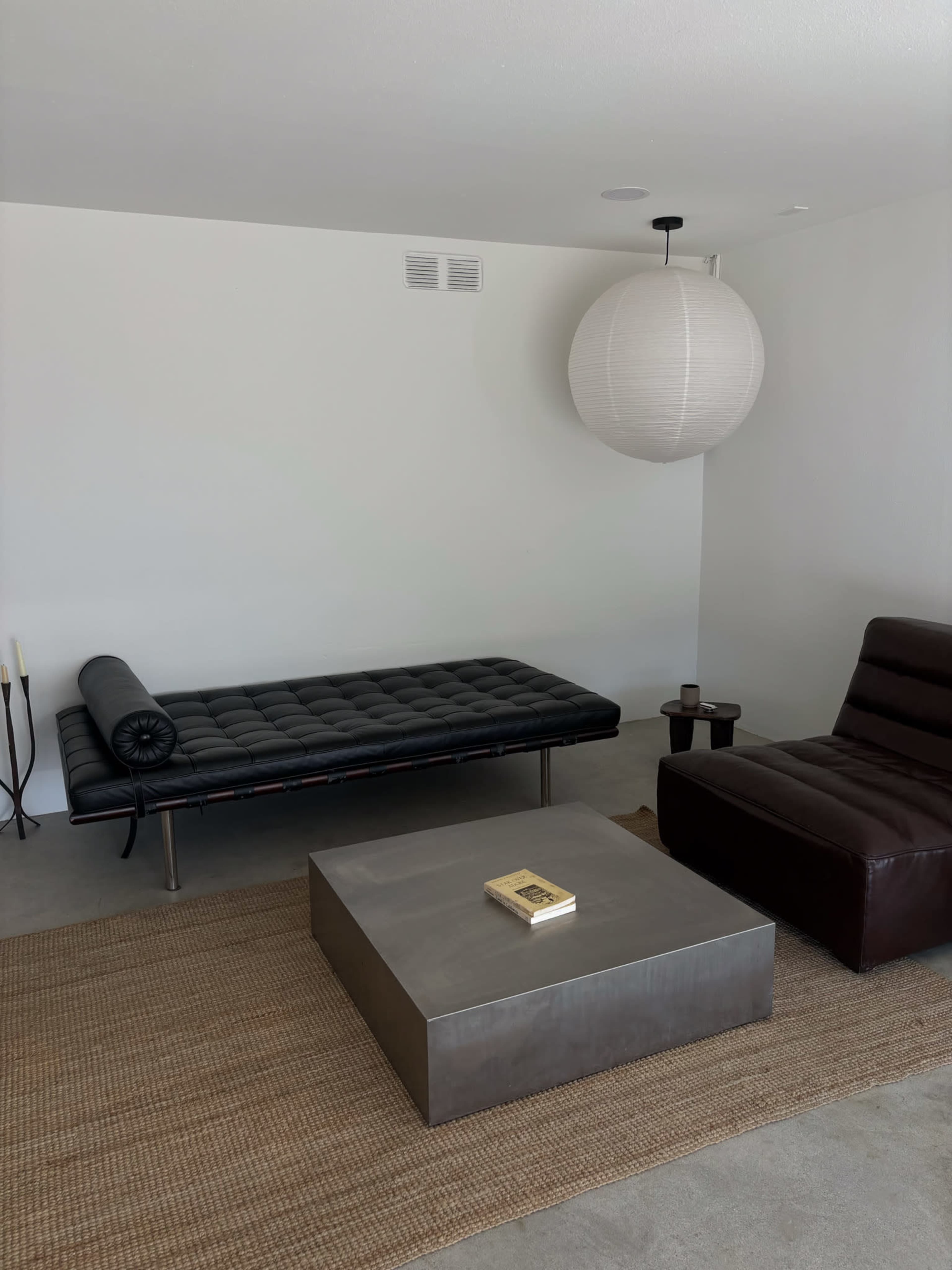The image shows a modern living room with a black leather daybed, a brown armchair, a metallic coffee table, and a round paper pendant lamp, all set on a neutral-colored floor and a woven rug.