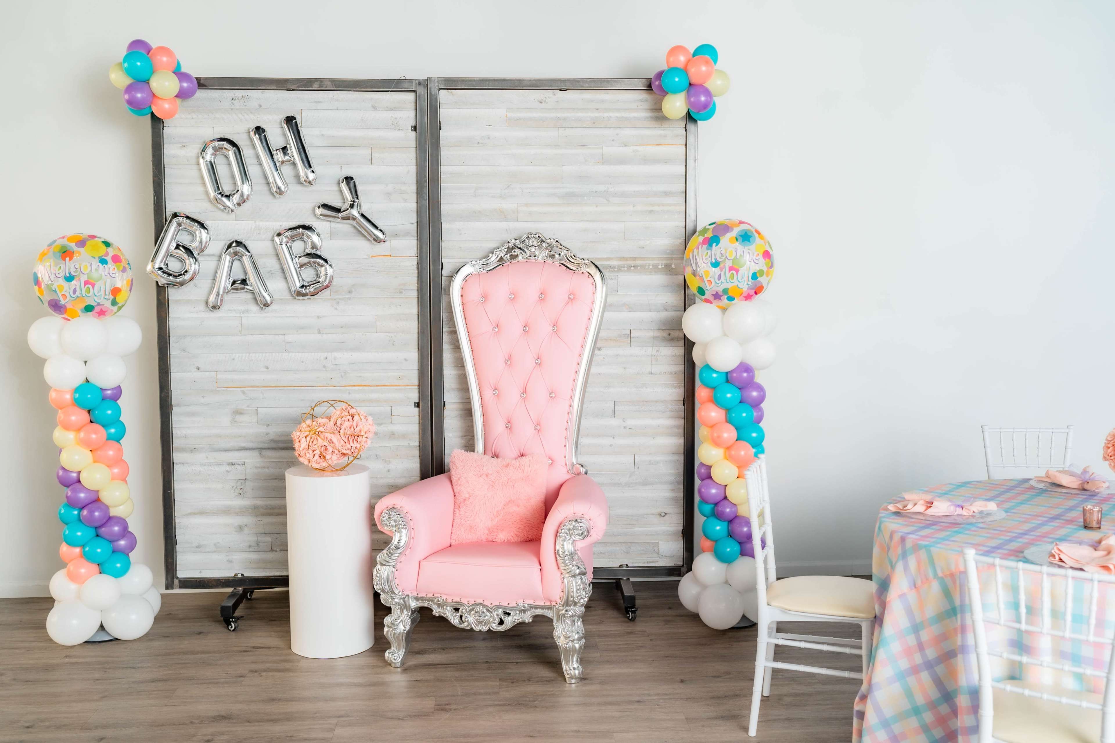 The scene features a decorative area with a pink throne chair, balloon columns, a sign saying "Oh Baby," and a table set with a plaid tablecloth.