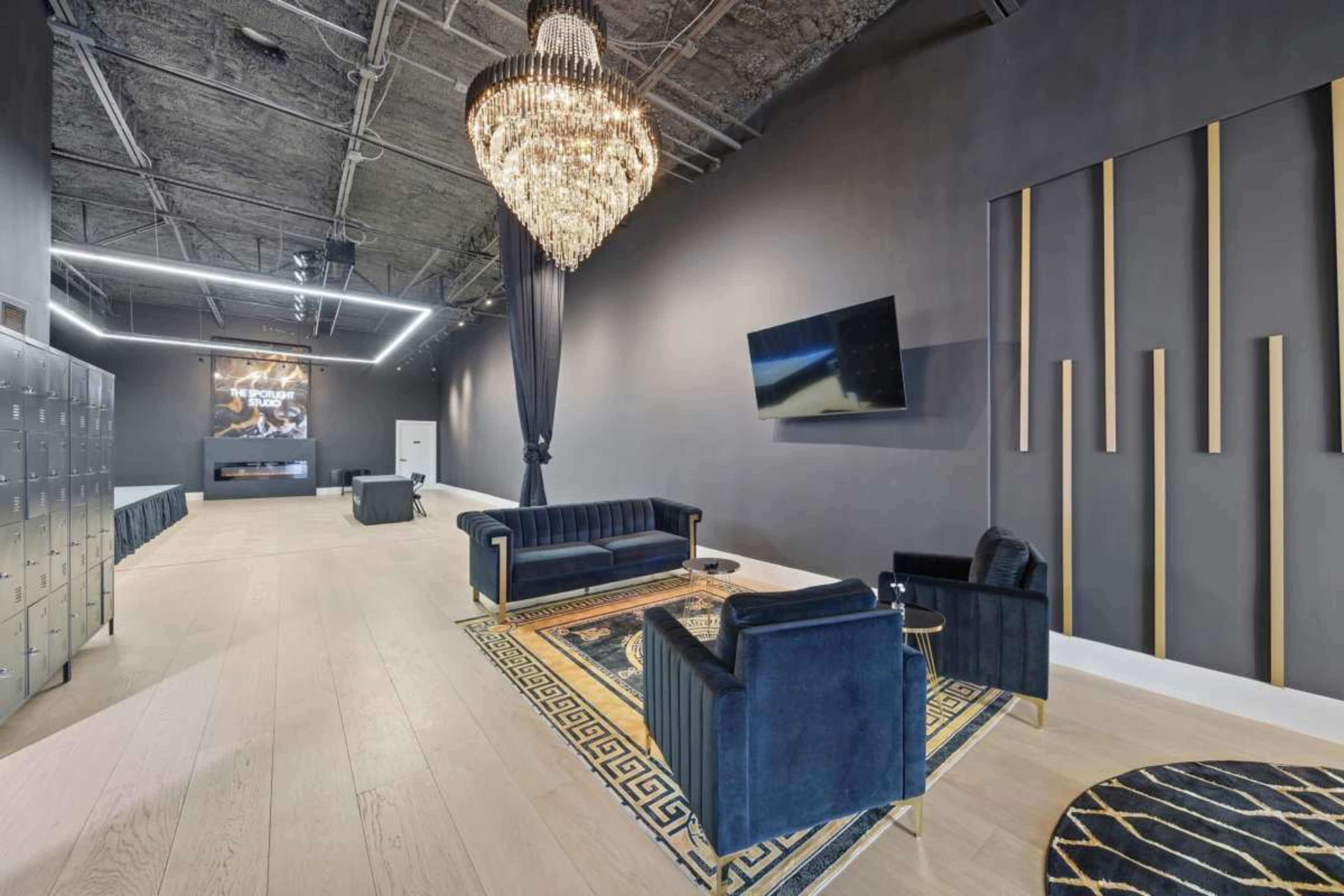 The image shows a modern lounge area with dark walls, a large chandelier, a flat-screen TV, and upholstered furniture, including a sofa and two chairs, set on a patterned rug.