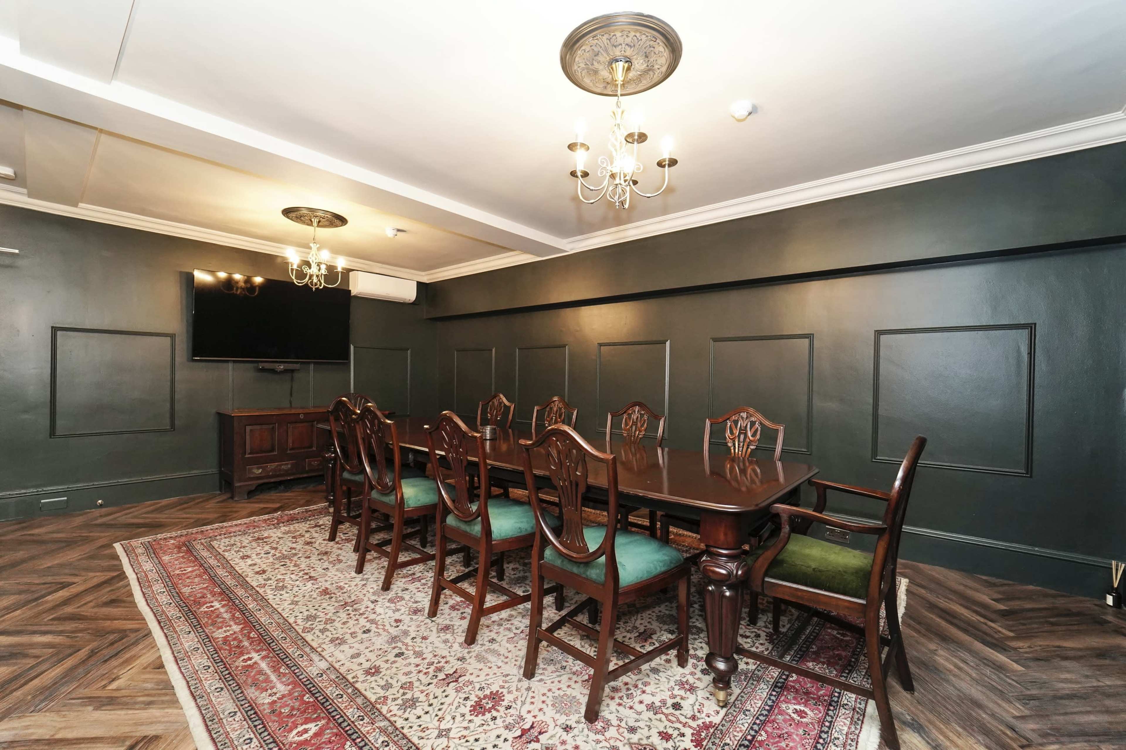 The image shows a dimly lit conference room featuring a long wooden table surrounded by upholstered chairs, set against a dark green wall with decorative paneling and a patterned rug on the floor.