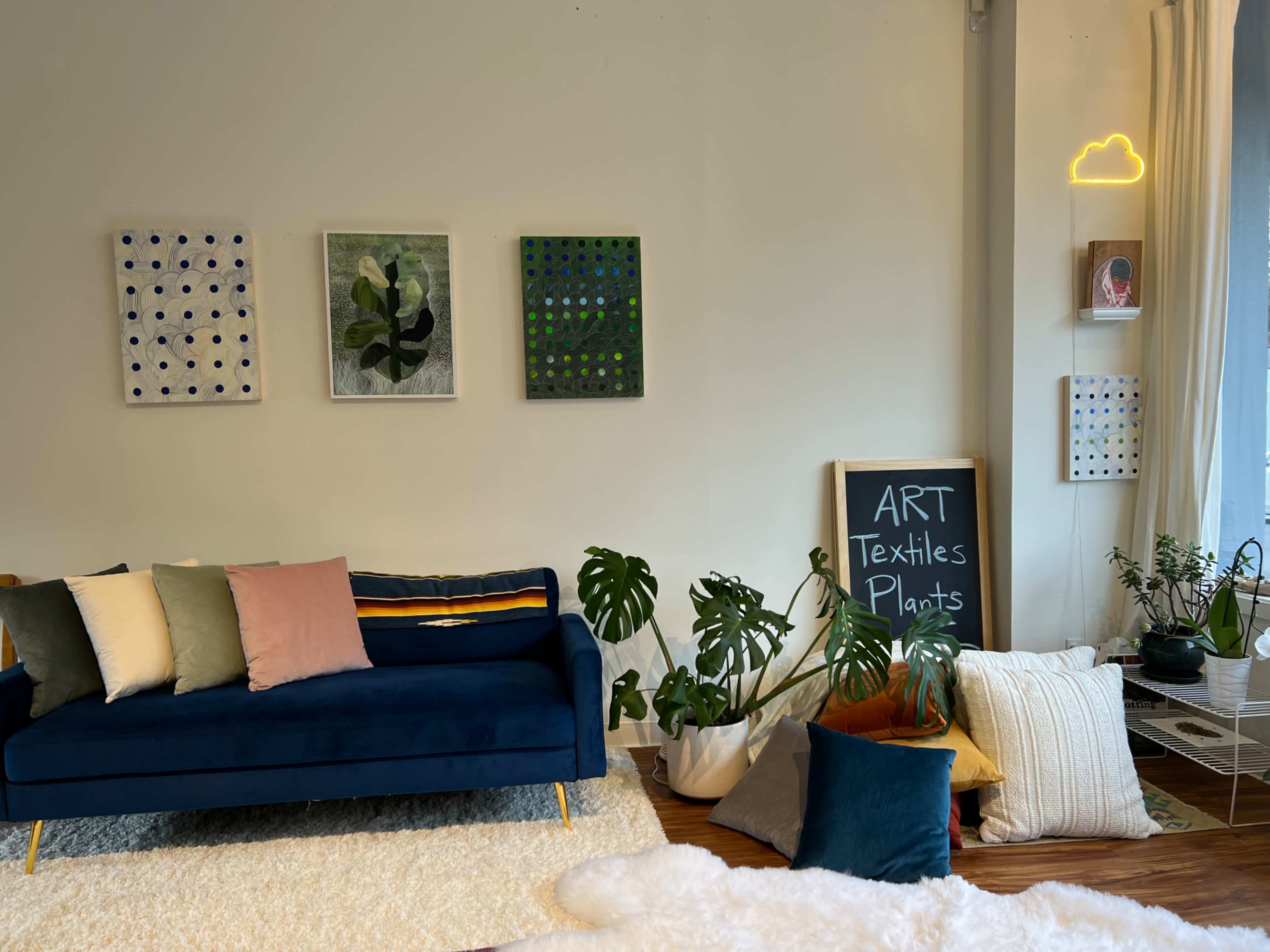 A cozy room features a dark blue sofa with various decorative pillows, a chalkboard listing "ART, Textiles, Plants," and several pieces of art on the walls, surrounded by indoor plants.