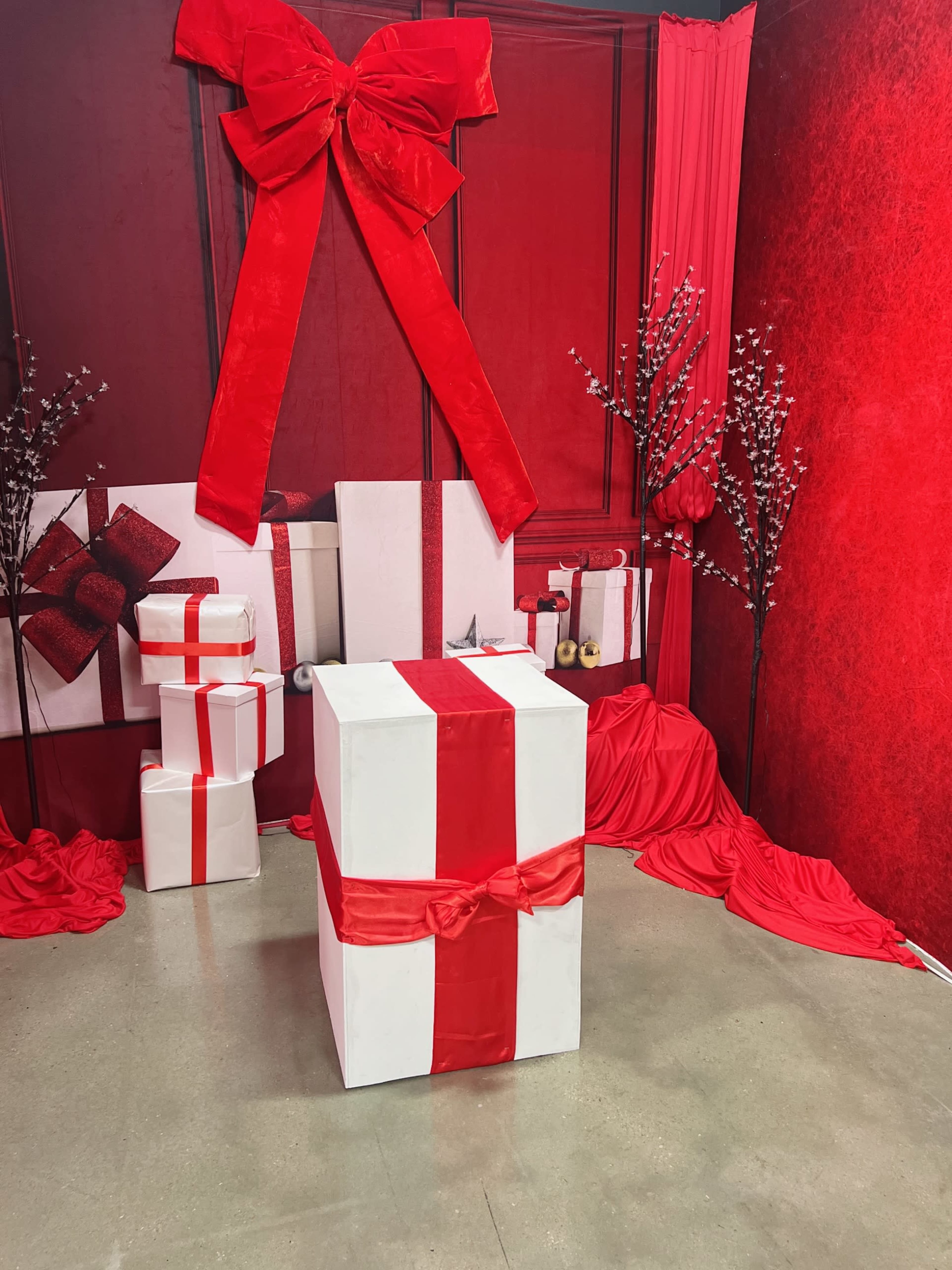 The scene features a large white gift box wrapped in a red ribbon, surrounded by smaller white and red gift boxes, red fabric draping, and decorative branches.