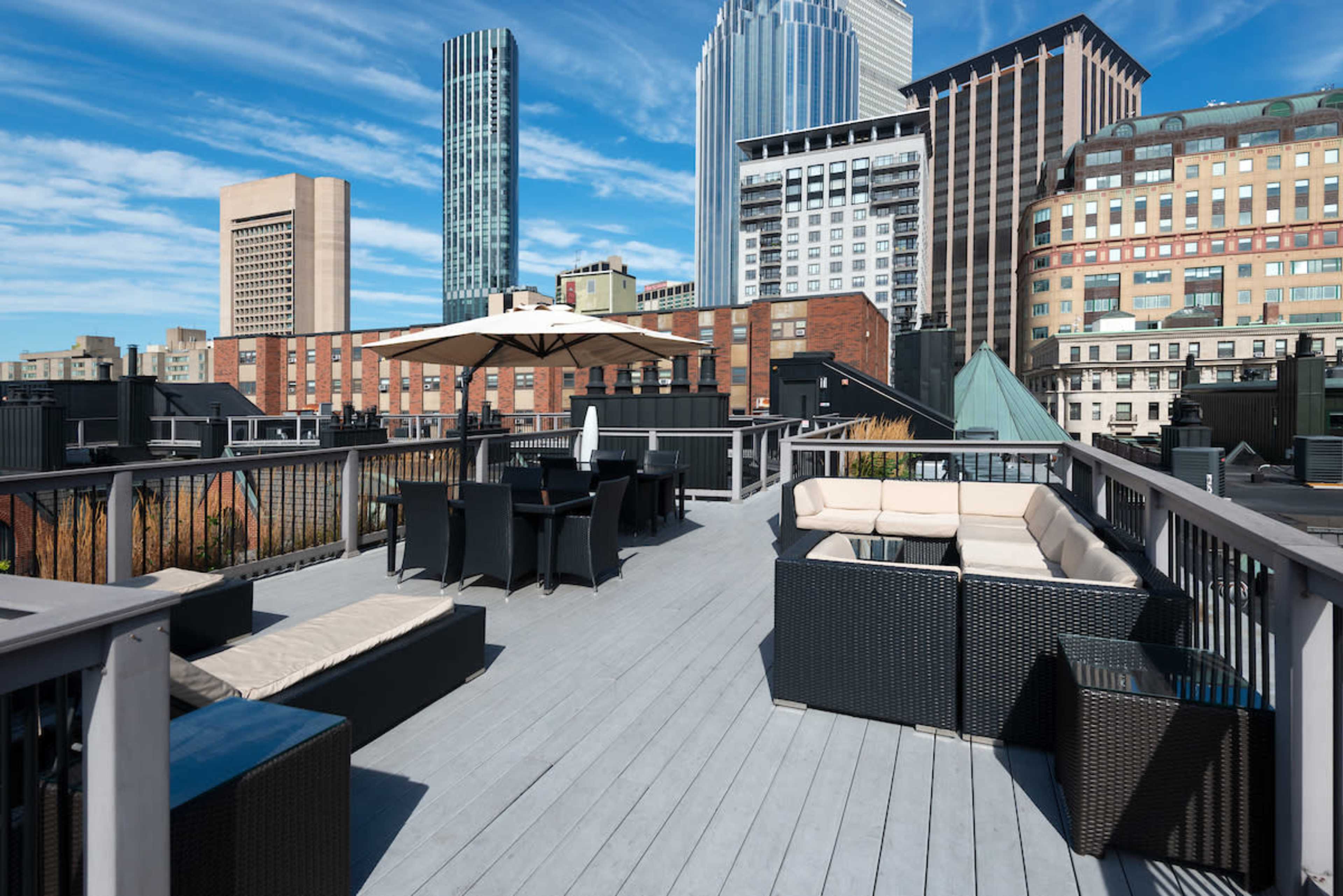 A rooftop terrace with seating areas, including lounge chairs and tables, overlooking a city skyline with tall buildings.