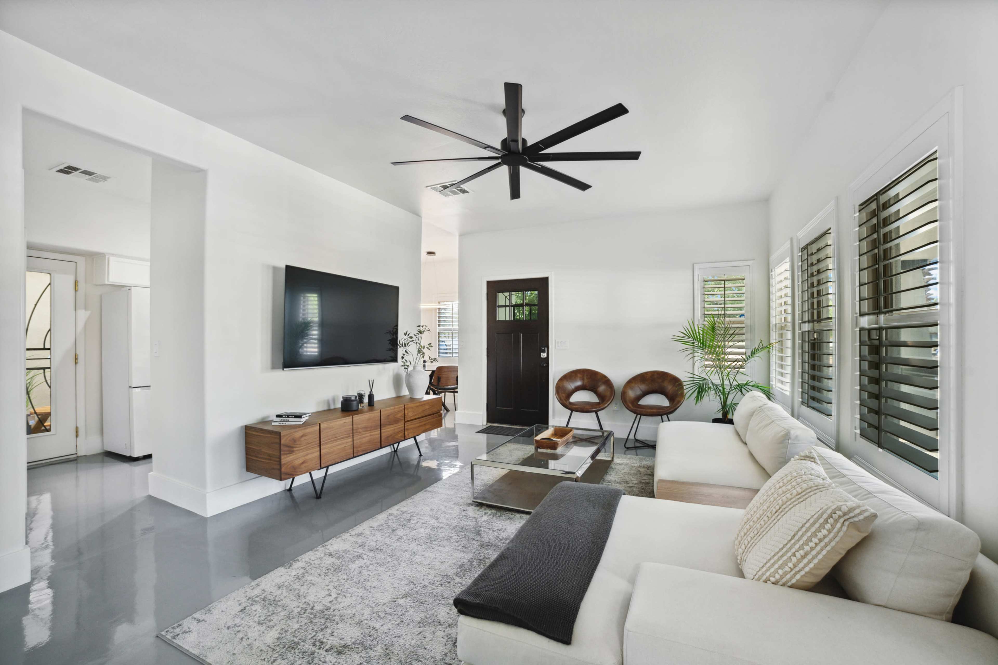 The living room features a white color scheme with a large television mounted on the wall, a sleek sofa, two brown chairs, and a potted plant near a black front door.