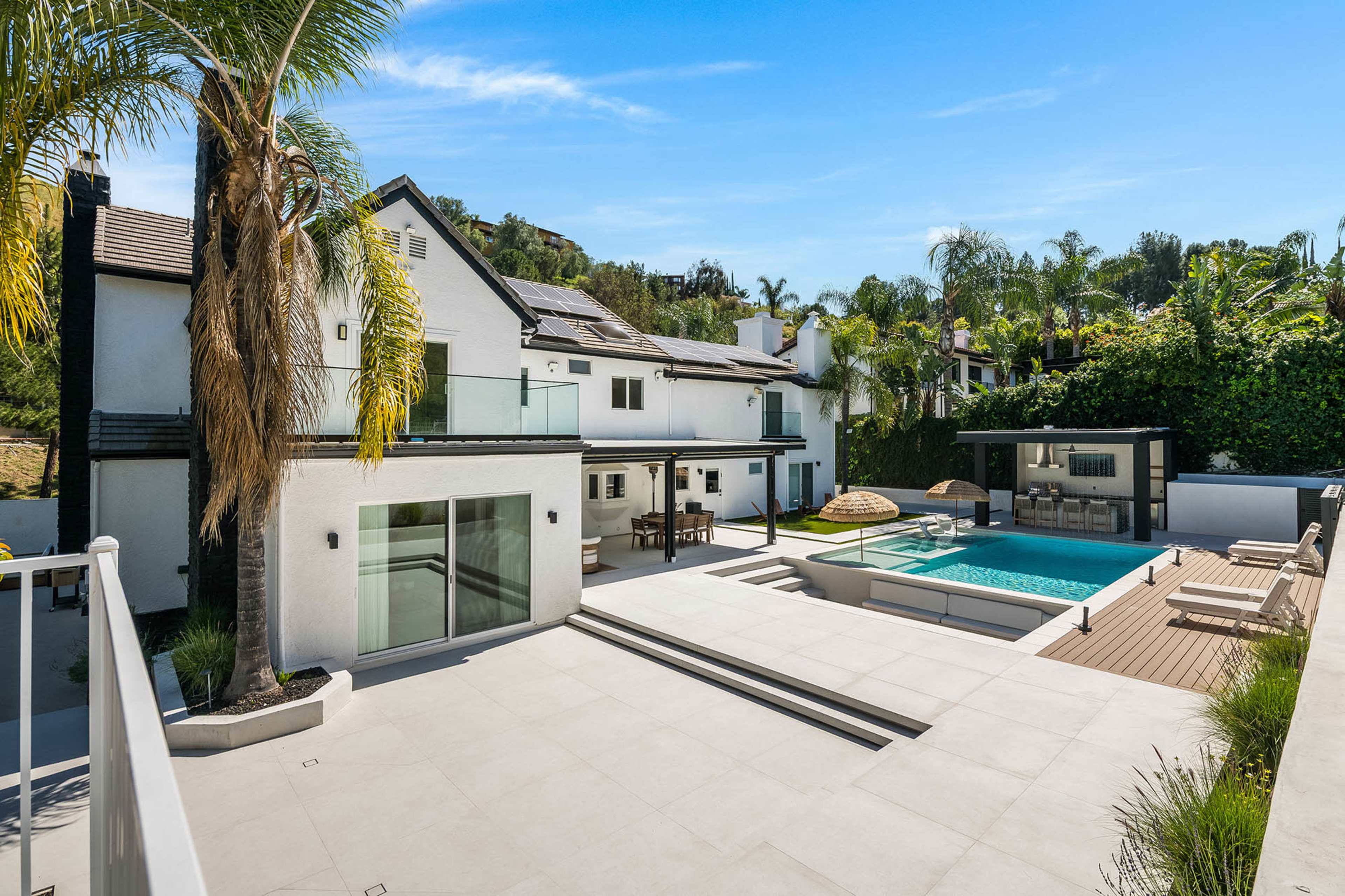 The image shows a modern house with a swimming pool, surrounded by palm trees and outdoor seating areas.