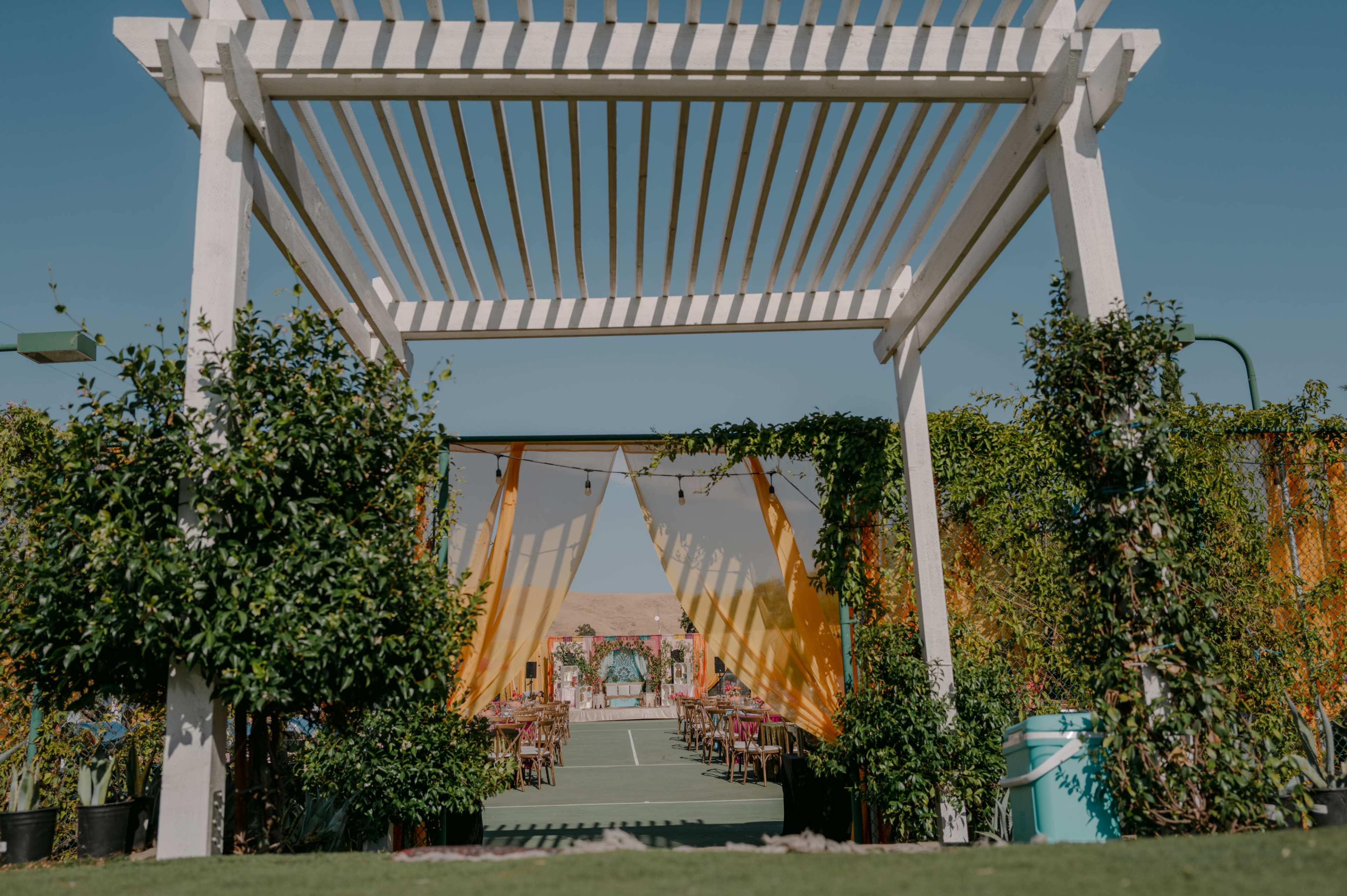 The image depicts an outdoor event space framed by a white pergola, with vibrant fabric drapes and greenery leading to a set of tables and chairs arranged for a gathering.