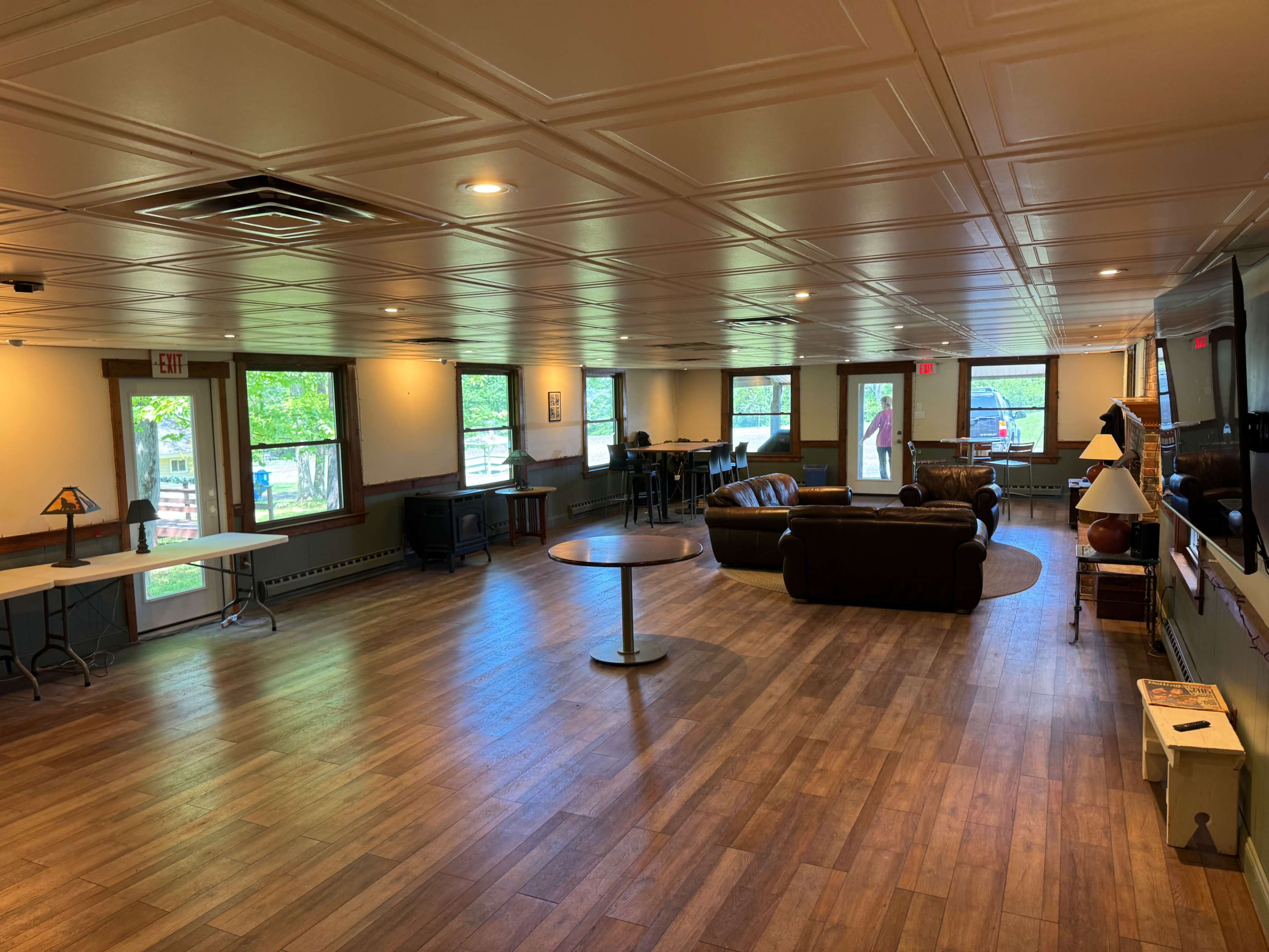 The image shows a spacious room with wooden flooring, multiple windows, and furniture including a couch, tables, and a television.