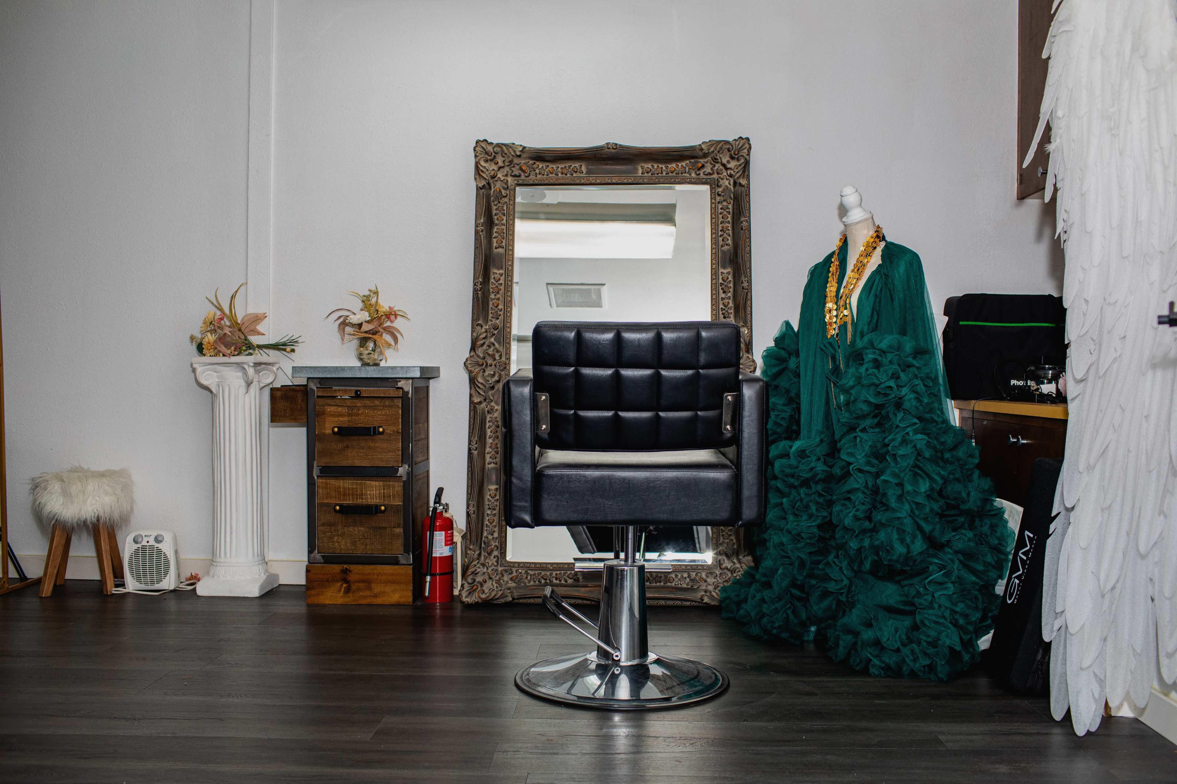 A black salon chair is positioned in front of a large ornate mirror in a hair salon, with a green ruffled dress displayed nearby.