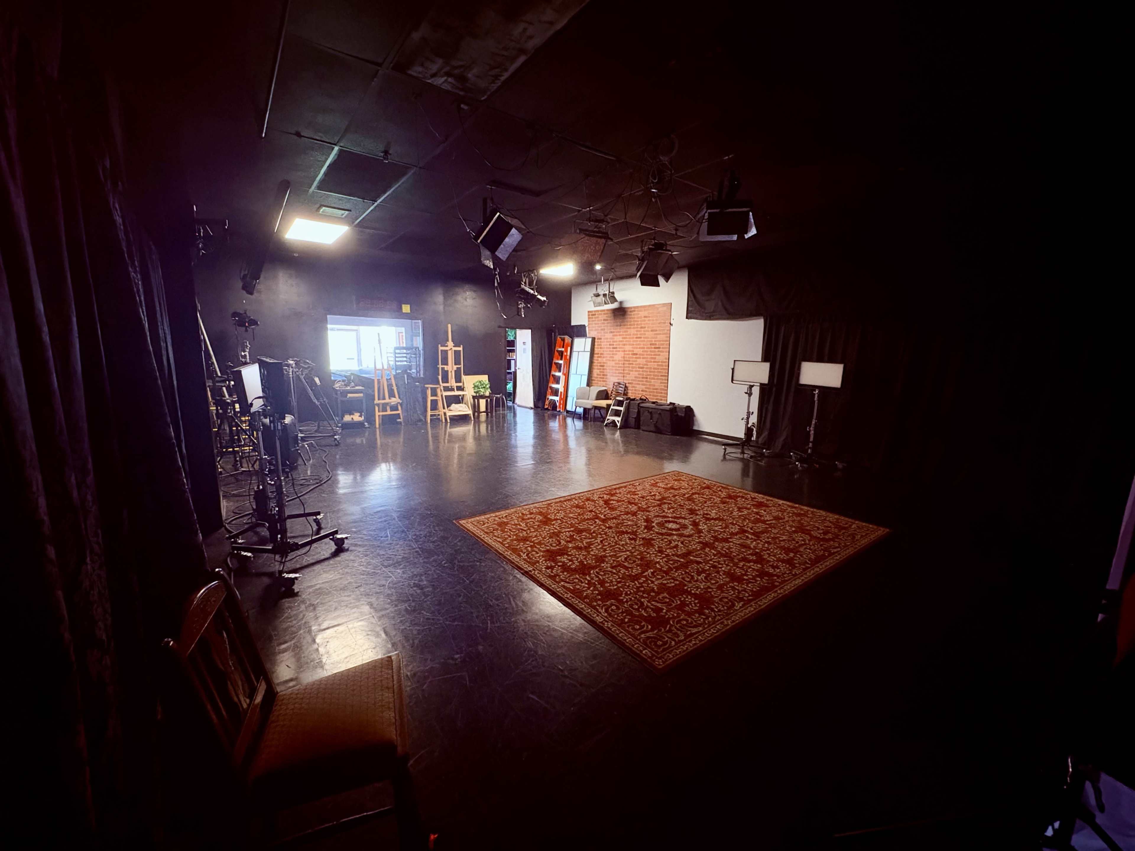 The image shows a spacious studio with a dark floor, several lighting setups, a large area rug in the center, and various props and furniture arranged along the sides.
