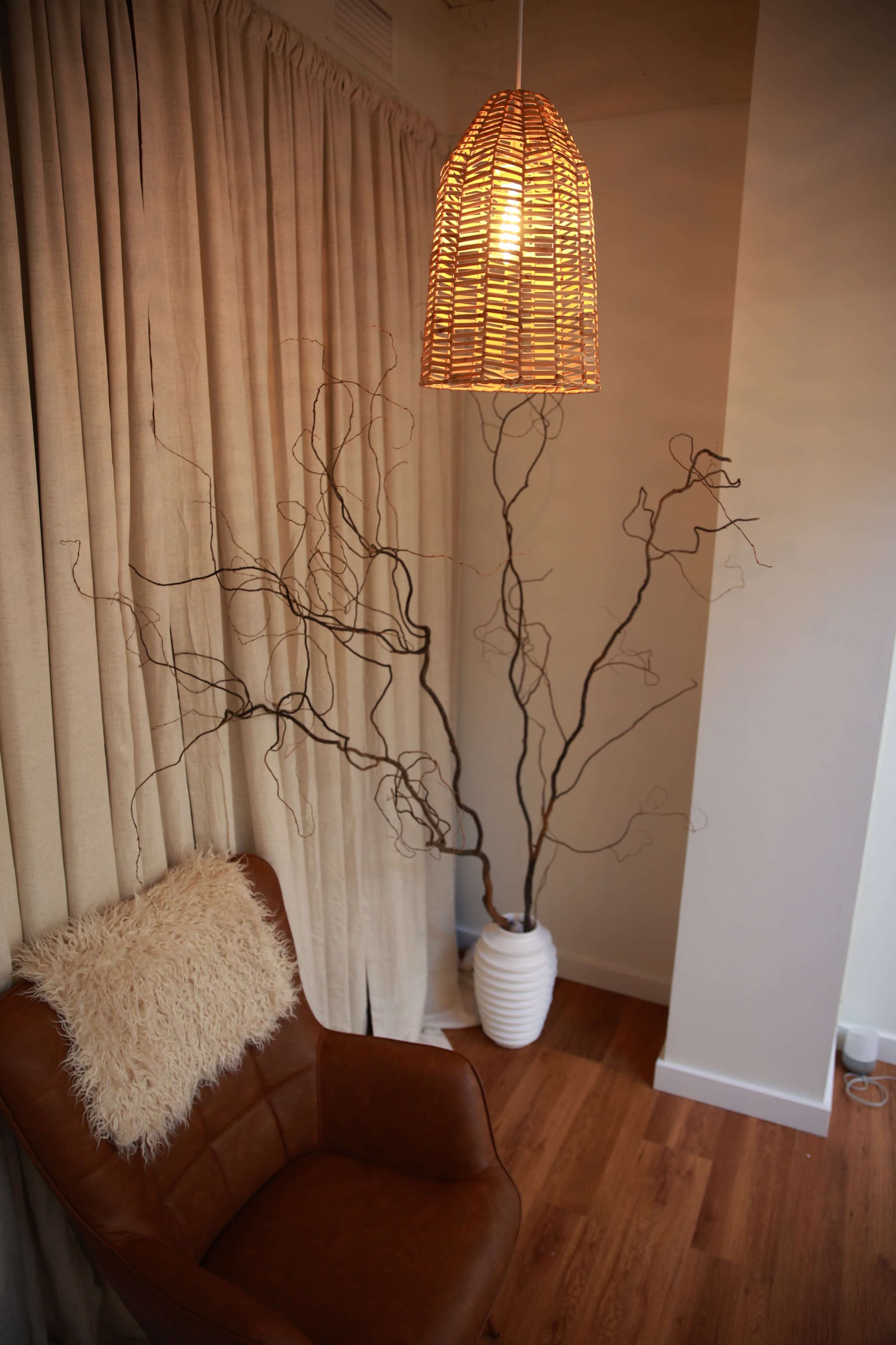 A comfortable brown chair sits beside a tall white vase containing bare branches, with a woven pendant light hanging above.