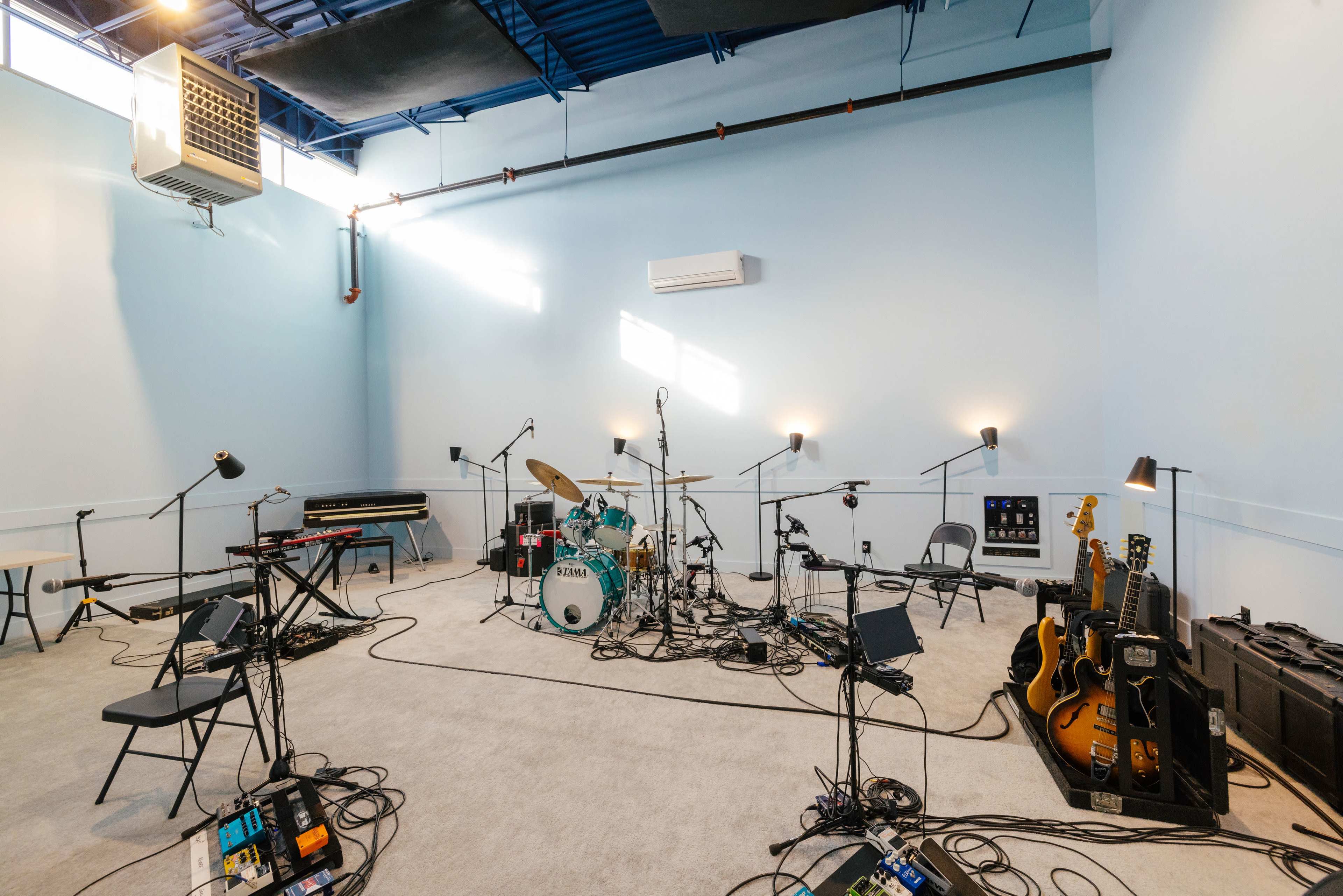 The image shows a music rehearsal space with various instruments, including a drum kit, keyboards, and guitars, arranged on an unadorned carpeted floor.