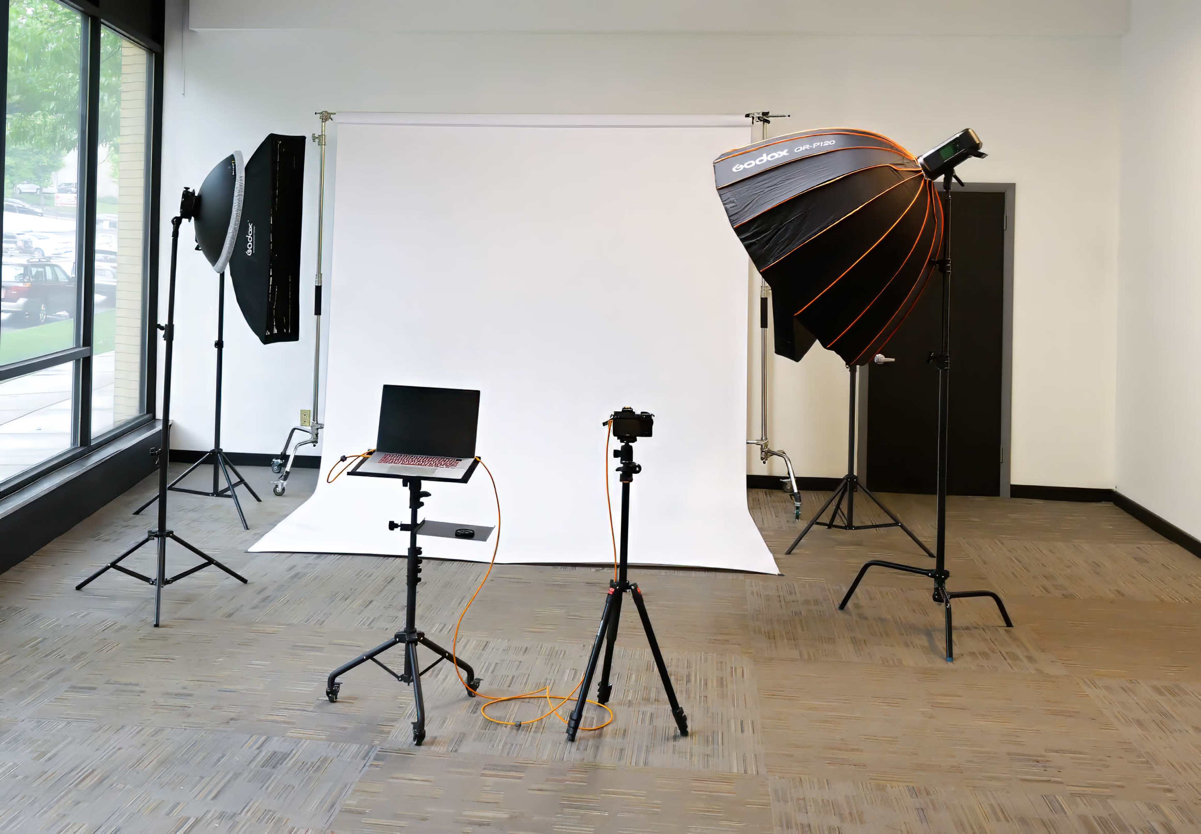 The image shows a photography studio setup with two light sources, a backdrop, a camera on a tripod, and a laptop on a stand.