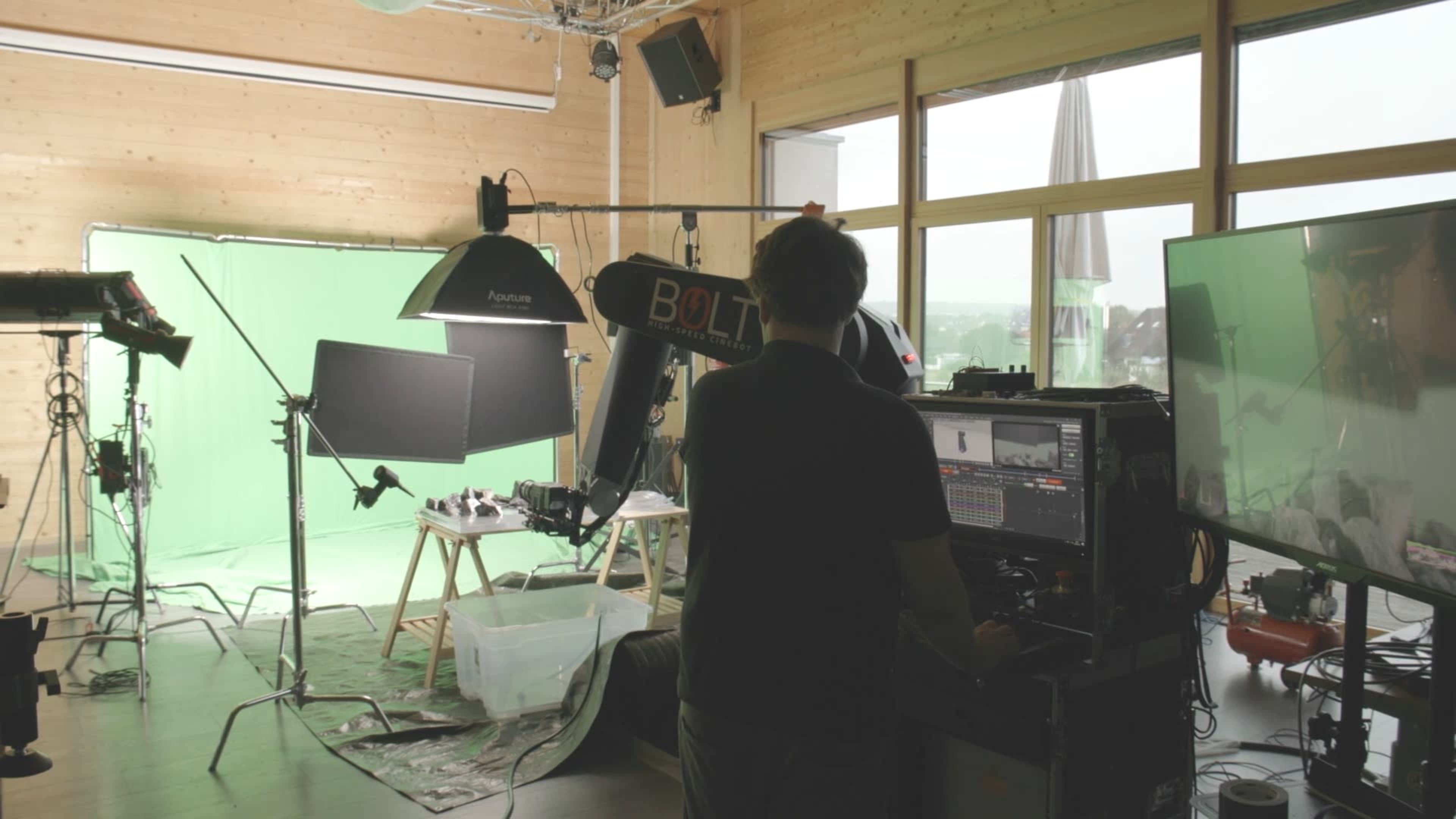 A film studio features a green screen setup, various lighting equipment, and a person operating a control panel in the foreground.