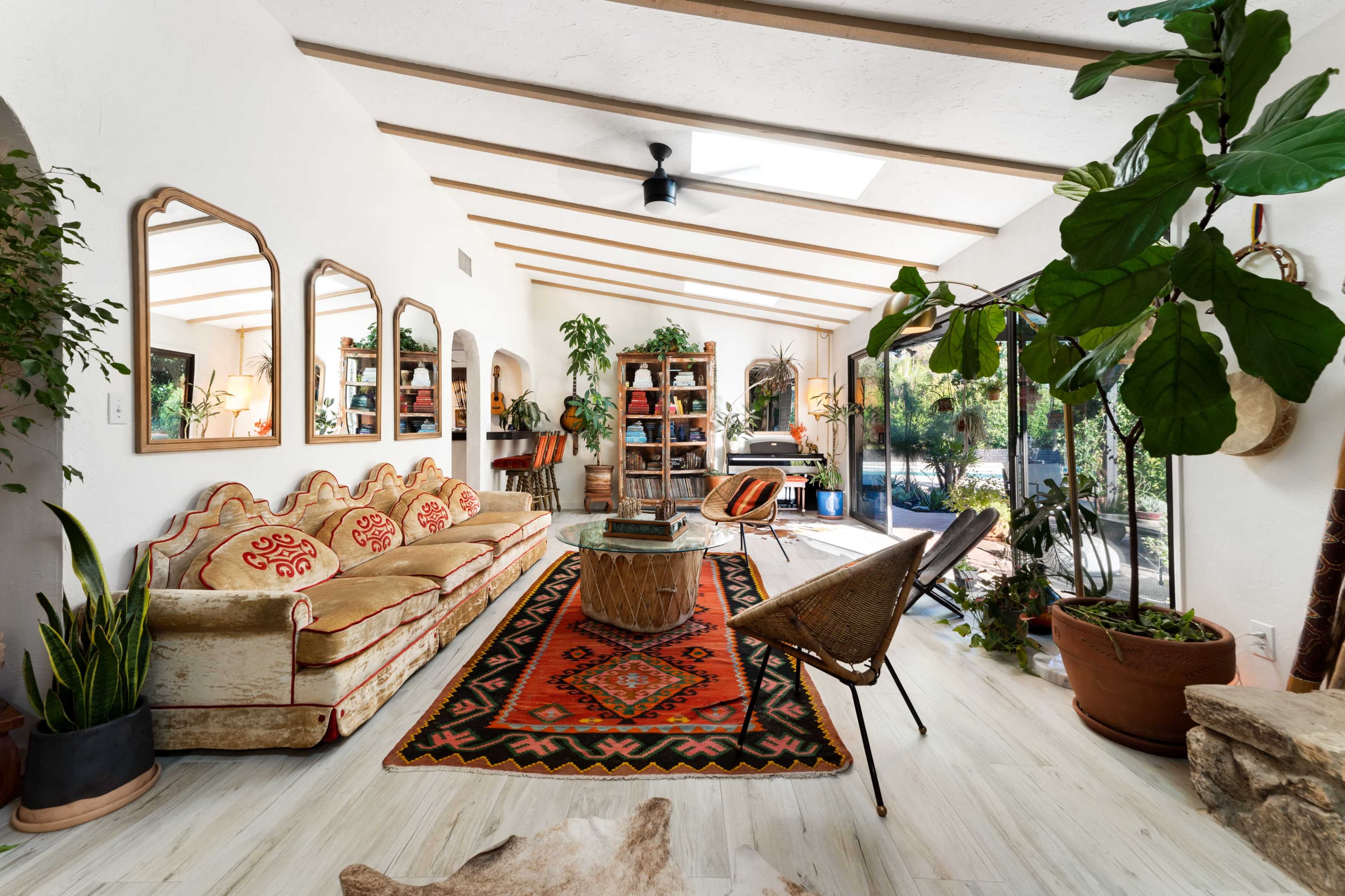 A bright, open living space featuring a vintage sofa, a patterned rug, plants, and large windows that provide natural light.