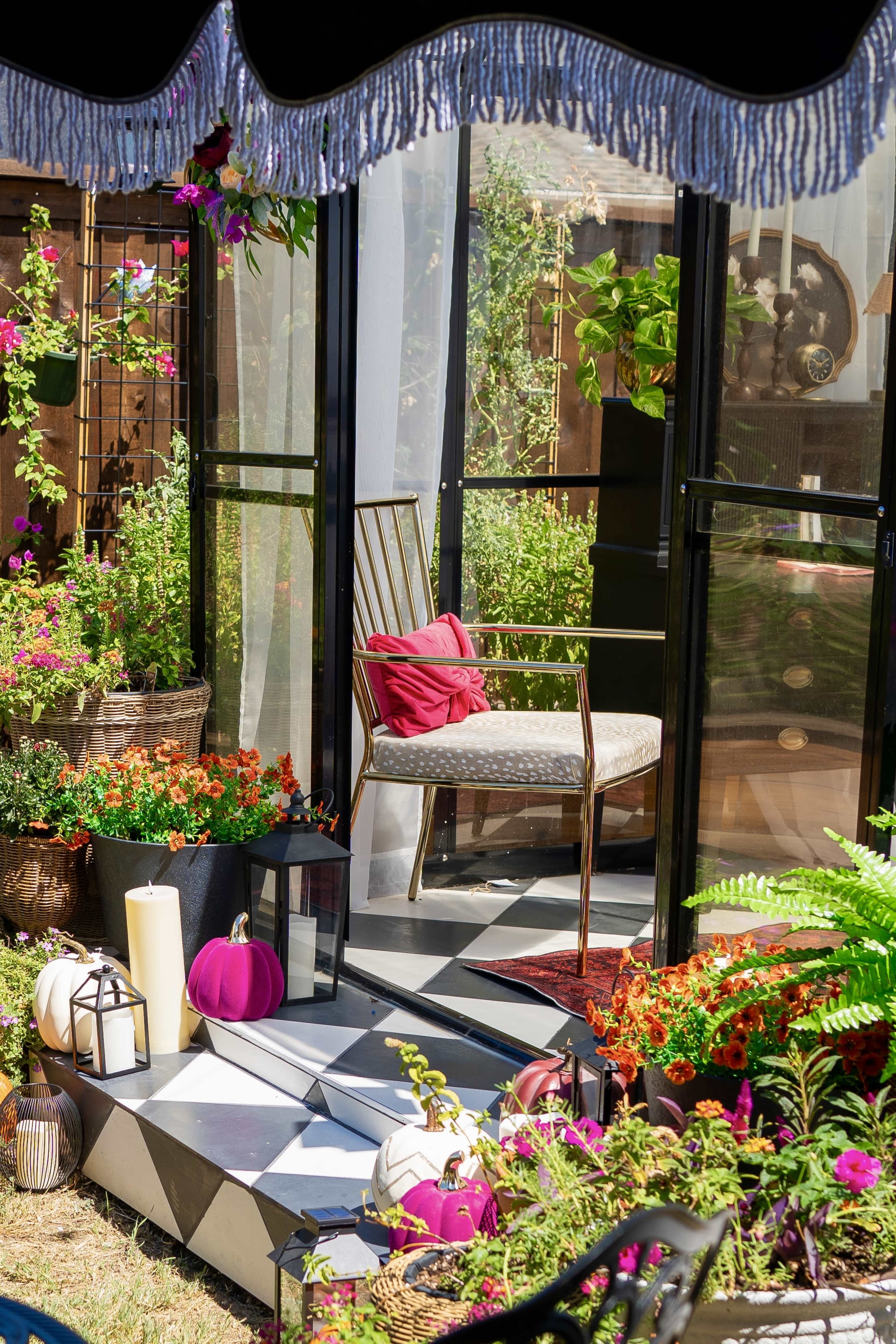 The scene features a bright outdoor space with colorful plants, a white upholstered chair with a red pillow, and various decorative pumpkins arranged among the greenery.