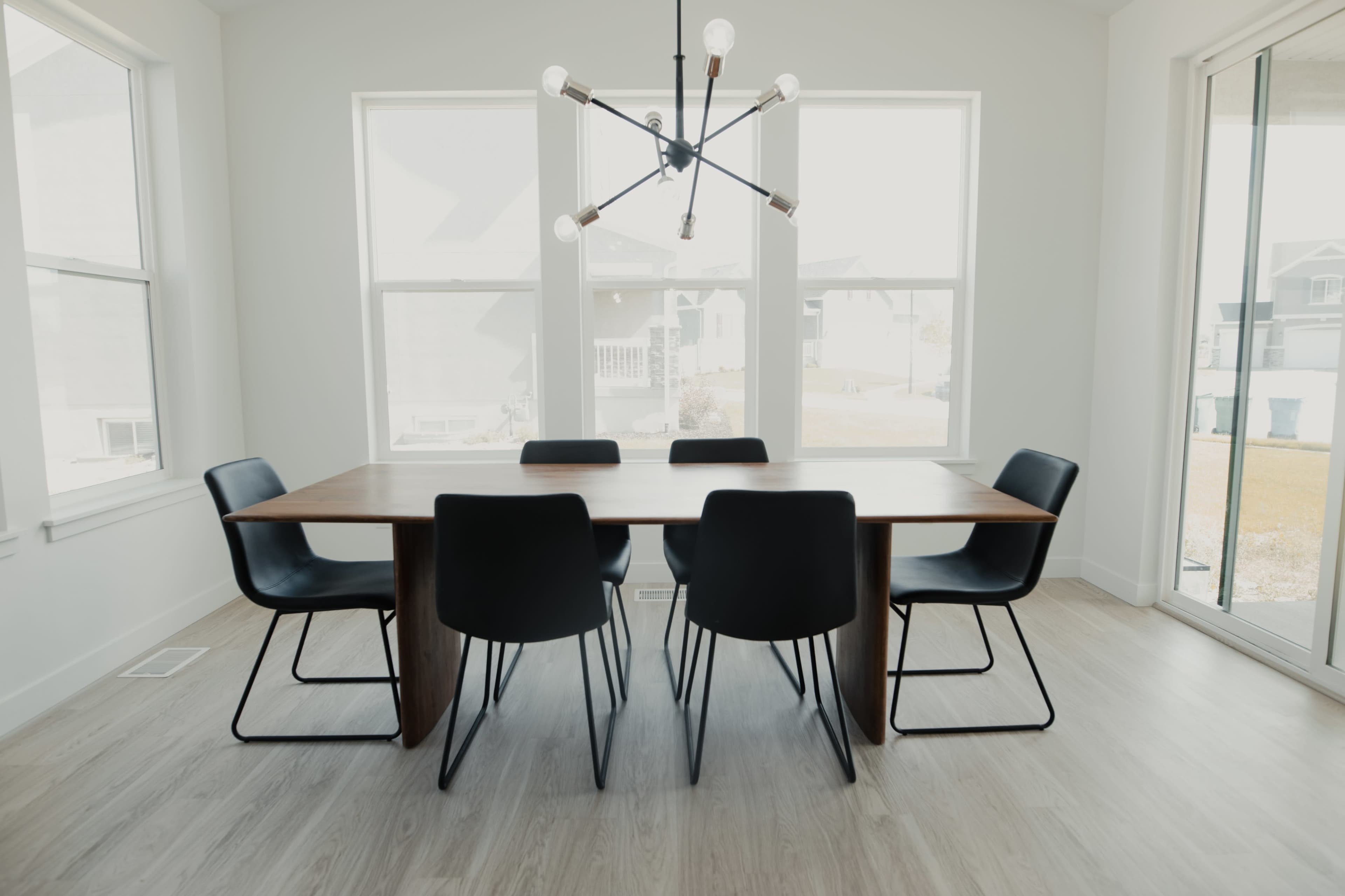 A modern dining room features a large wooden table surrounded by black chairs, with a unique light fixture above and large windows on two sides.