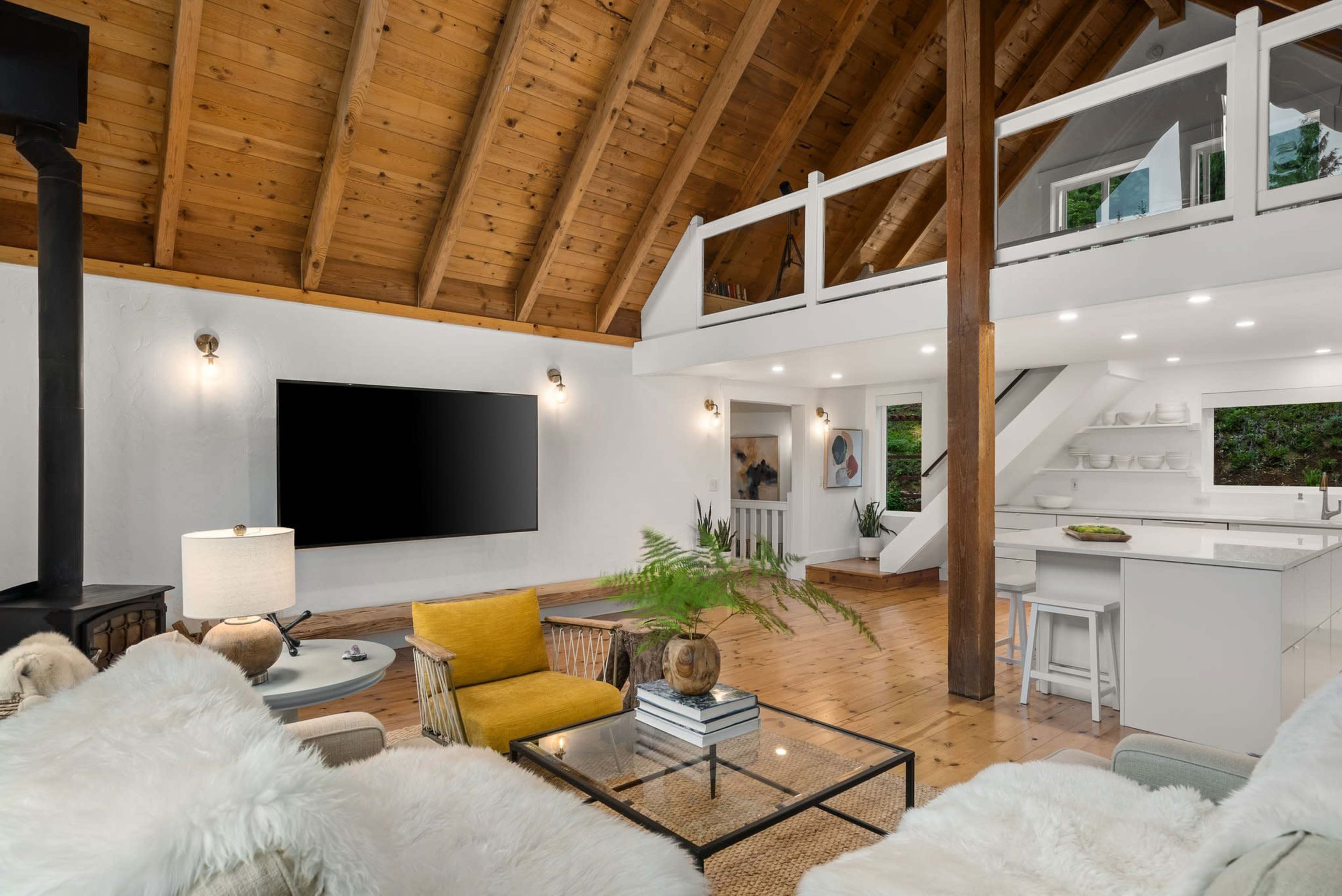 A modern living area features vaulted wooden ceilings, a large flat-screen TV, and a kitchen with white cabinetry and a staircase leading to a loft space.