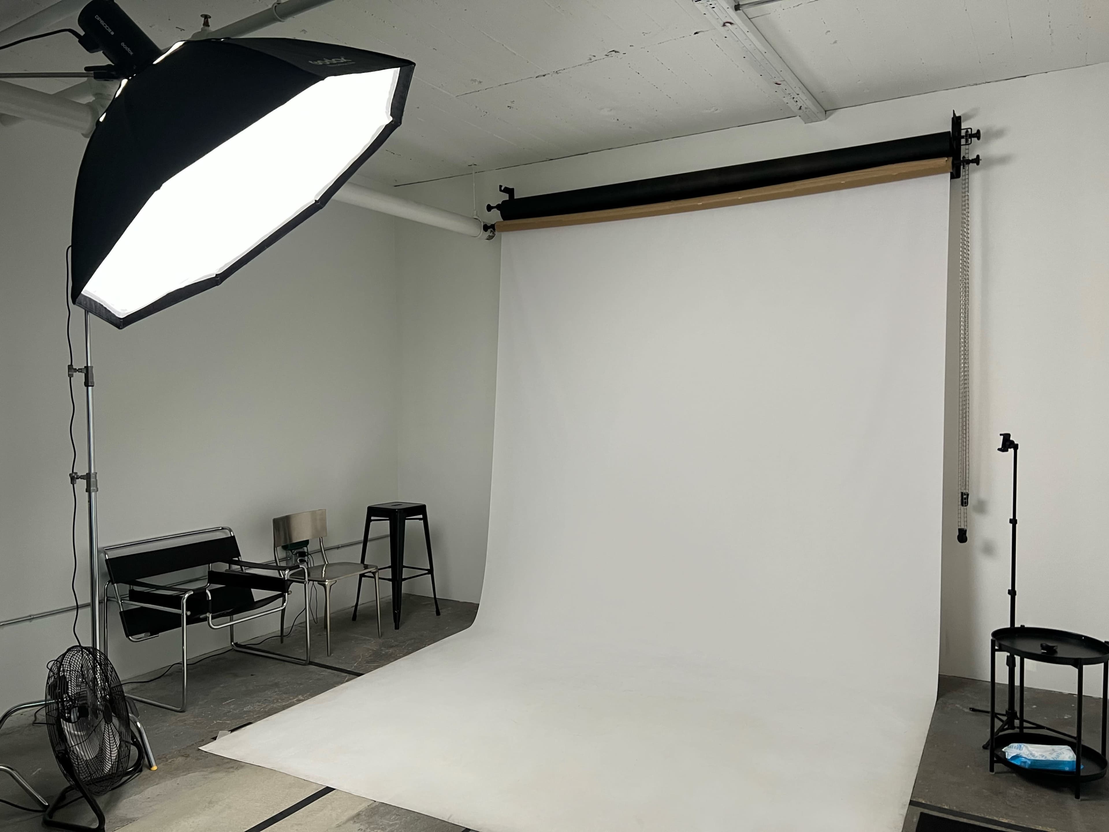 The image depicts a photography studio featuring a blank white backdrop, a large softbox light, a chair, a stool, and a fan.