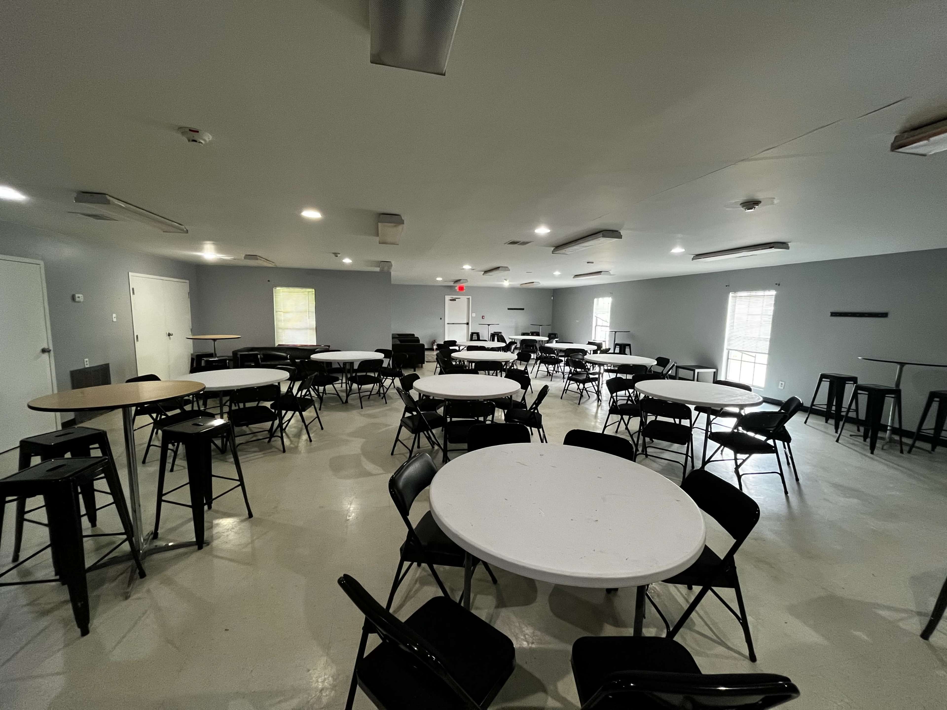 The image shows a spacious room layout featuring several round and square tables surrounded by black chairs, with bright overhead lighting and multiple windows.