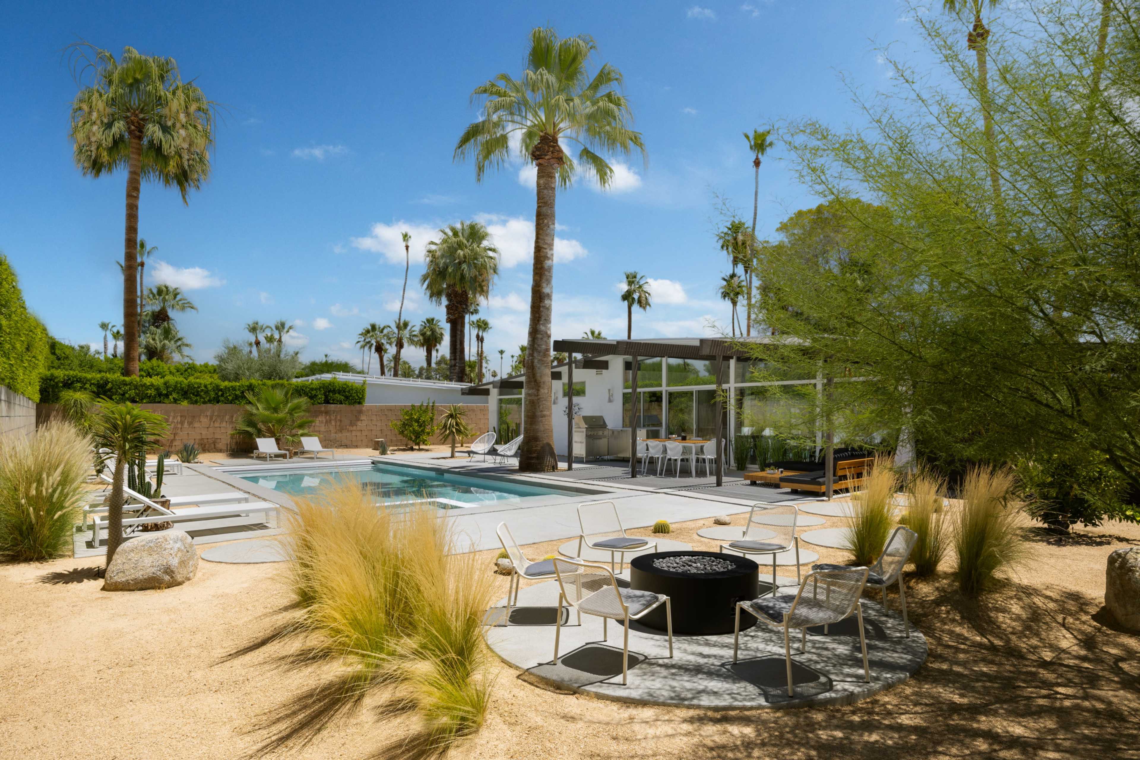 The image depicts a modern backyard with a swimming pool surrounded by palm trees, lounge chairs, and a seating area featuring a fire pit.