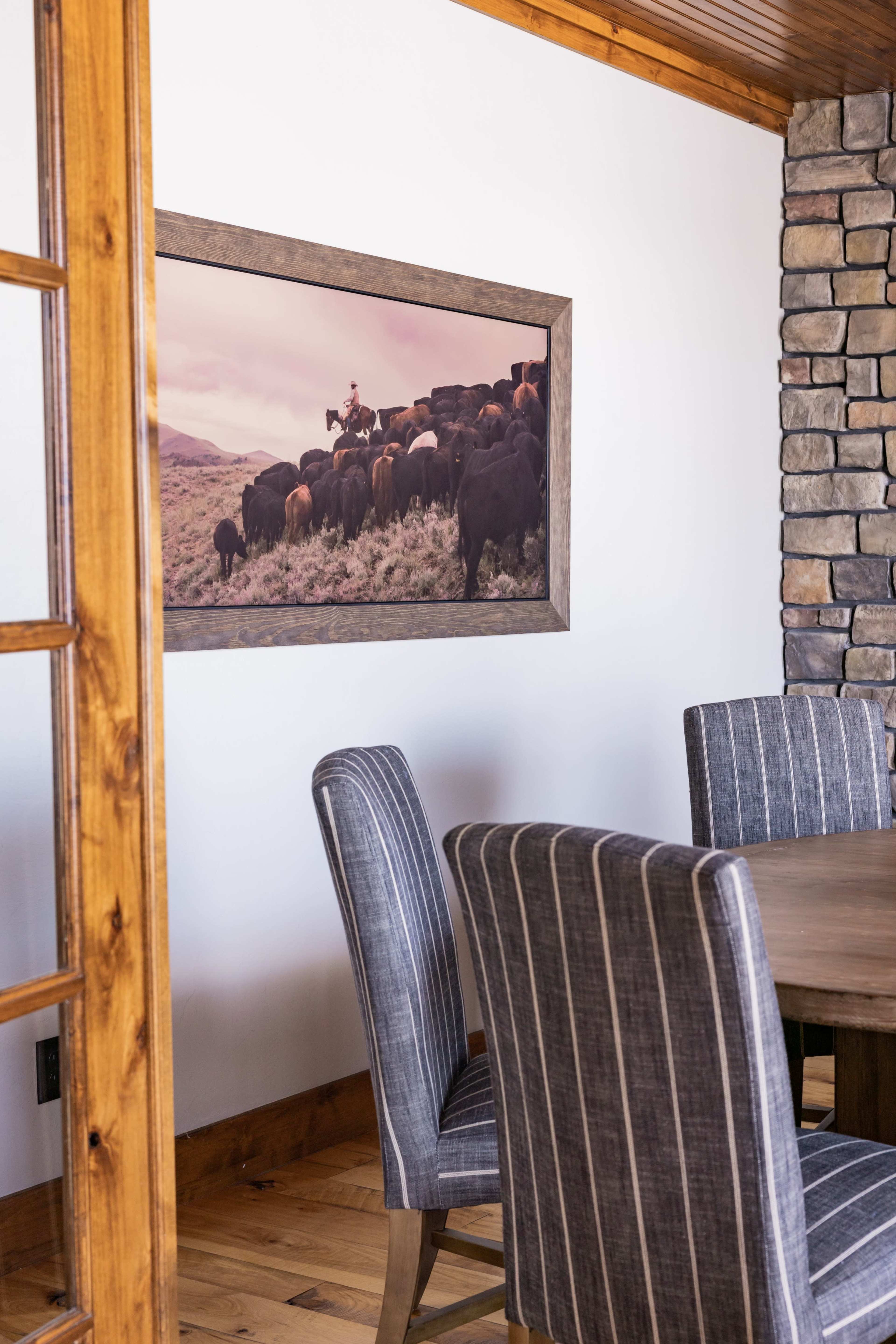 A rustic dining area features a wooden table surrounded by striped chairs, with a large framed photograph of cattle mounted on the wall.