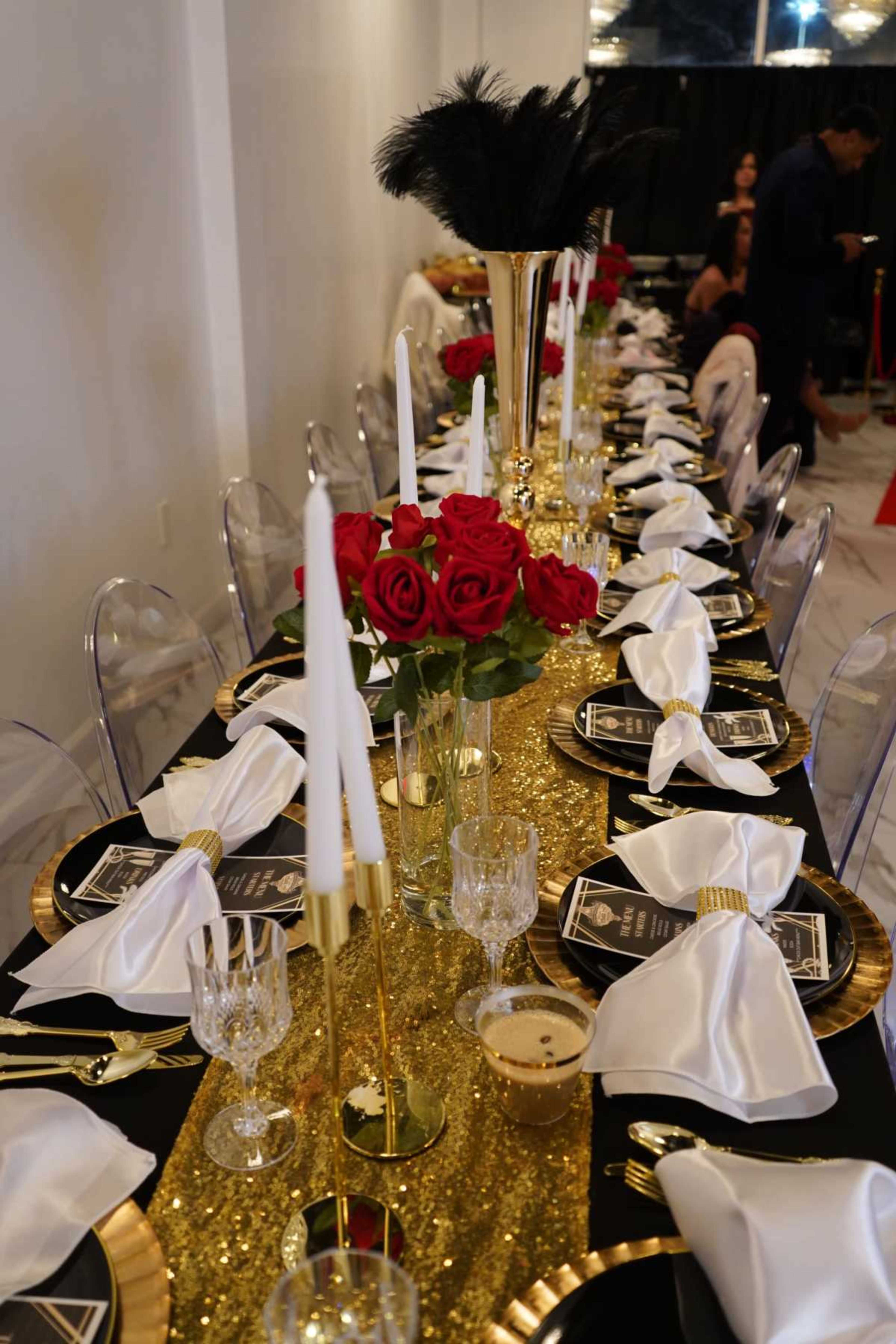 A long dining table is elegantly set with white napkins, black plates, and gold decorations, featuring red roses in a vase and tall candle holders.