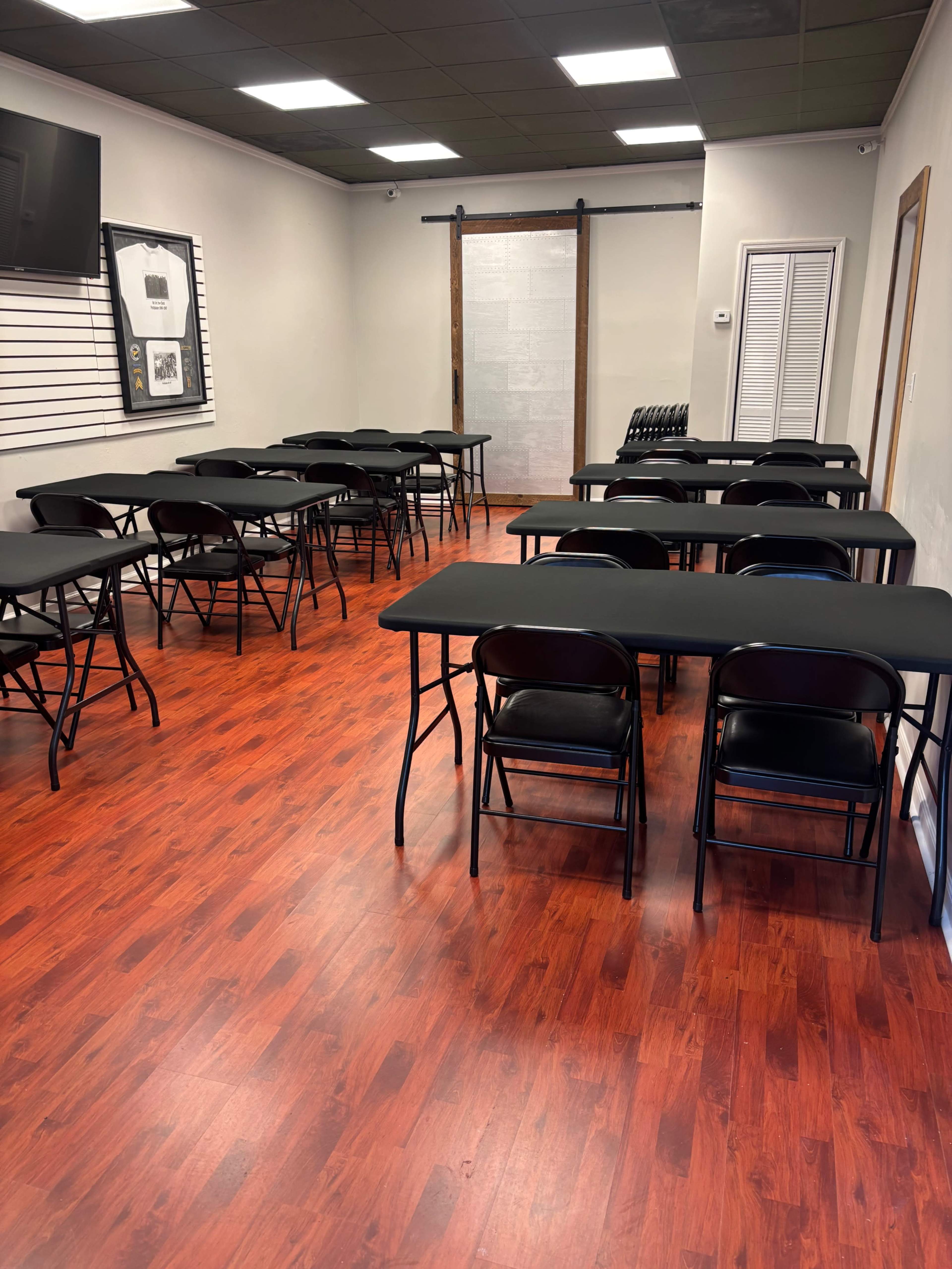 The room features several rows of black folding tables and chairs arranged on a polished wooden floor, with a sliding barn door and wall-mounted television.