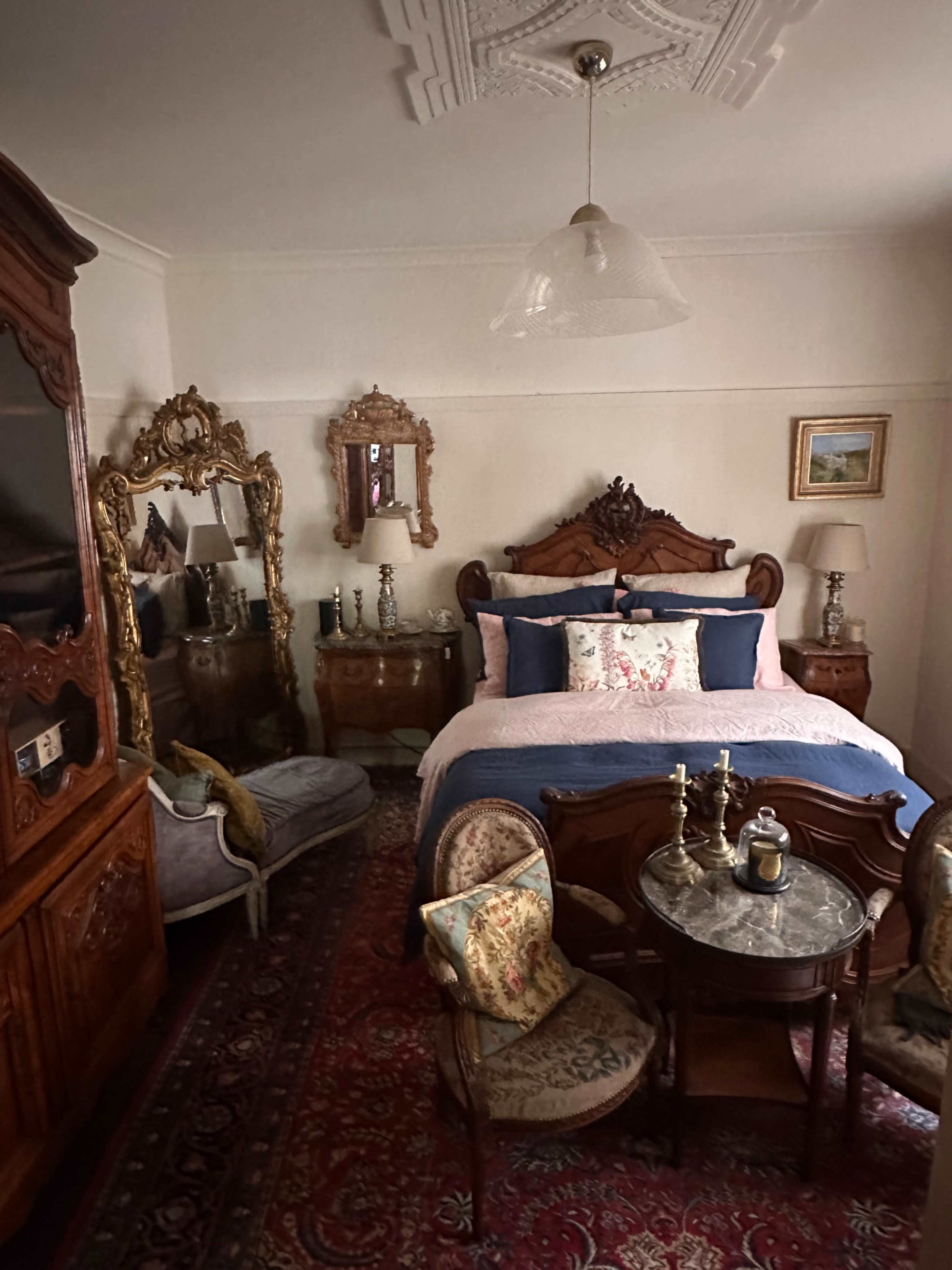 The image shows a vintage-style bedroom featuring a large bed with decorative pillows, an ornate wooden cabinet, and a small round table accompanied by two chairs.