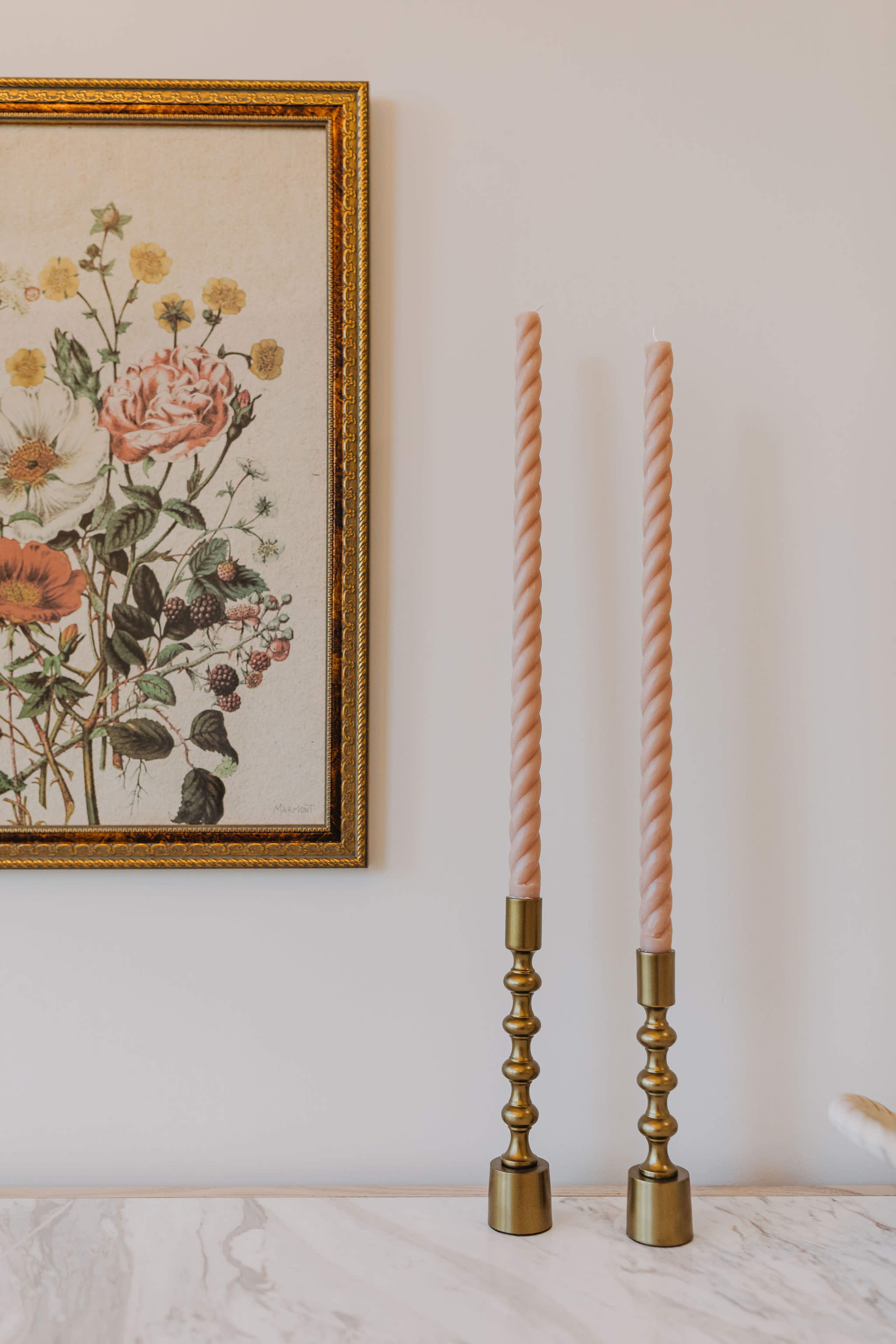 The image shows two twisted pink candles in brass holders beside a vintage floral print framed in gold on a wall.