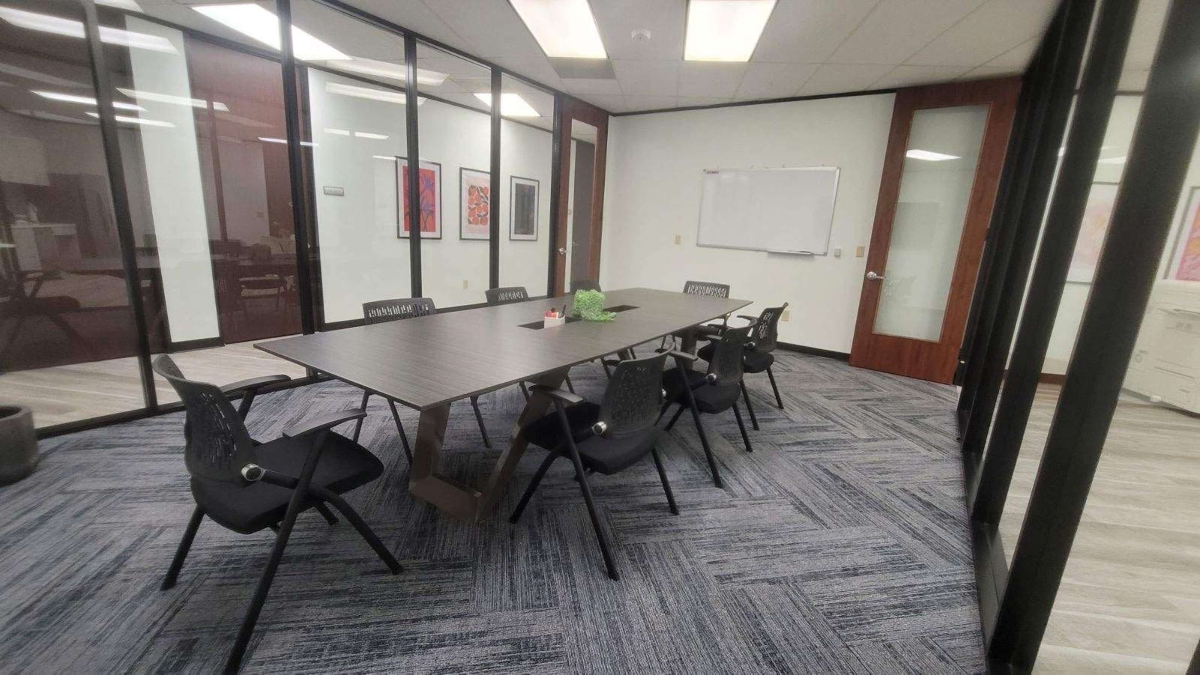 A modern conference room features a long table surrounded by black chairs, with glass walls and a whiteboard in the background.