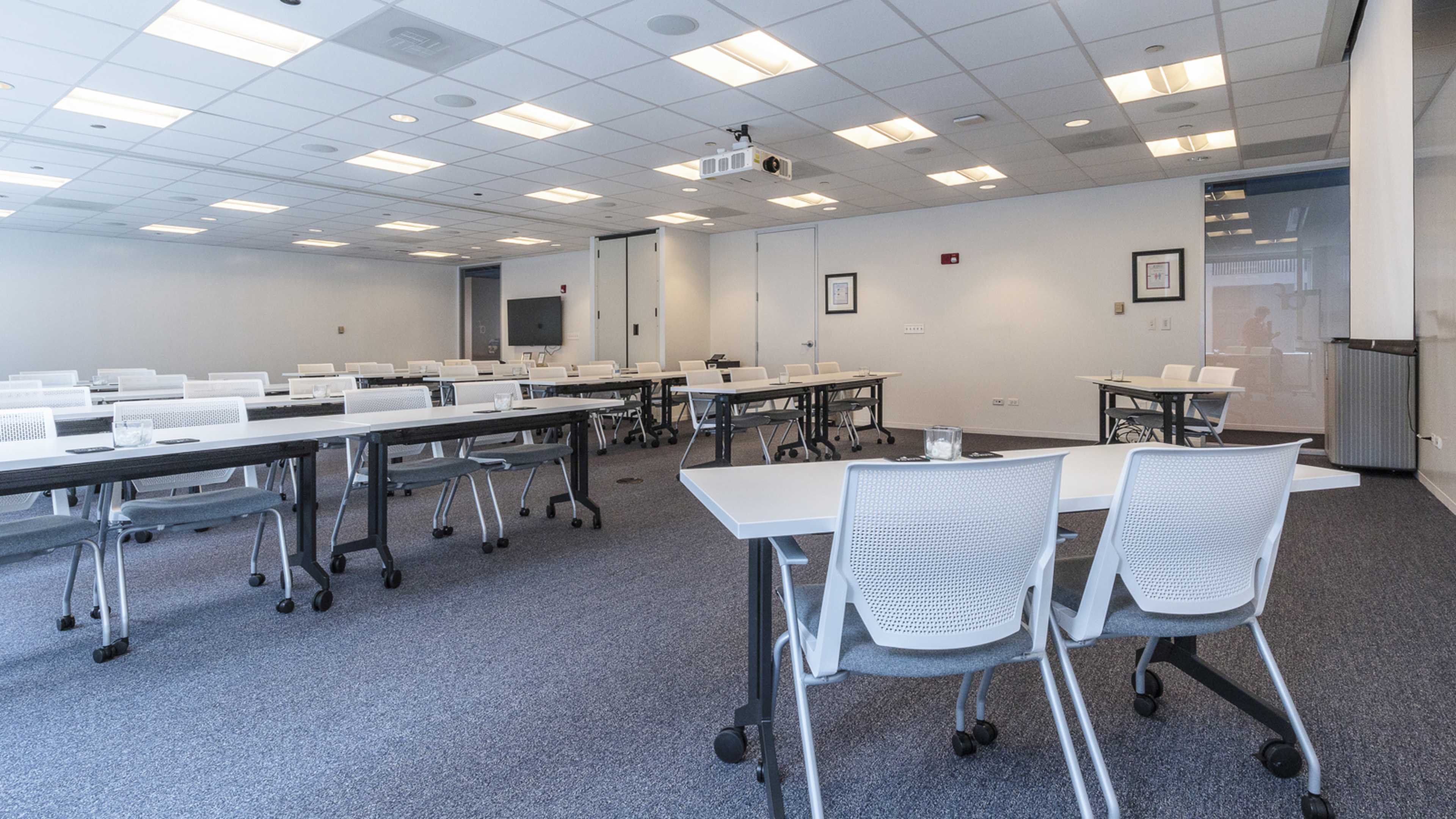 A spacious conference room is set with rows of tables and chairs, equipped with a projector and whiteboard.