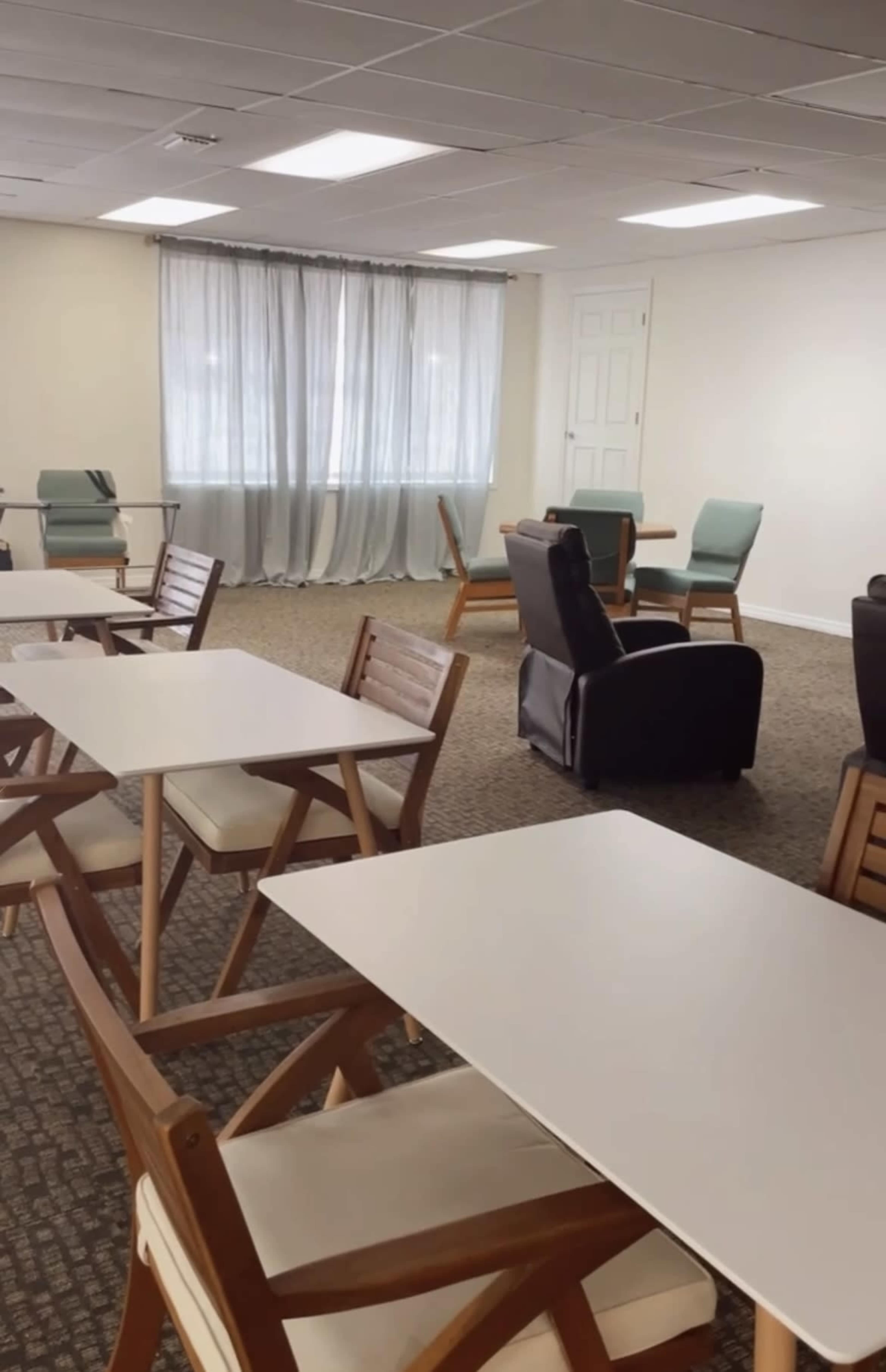 A spacious room with several tables and chairs arranged in a seating area, along with large windows covered by light curtains.