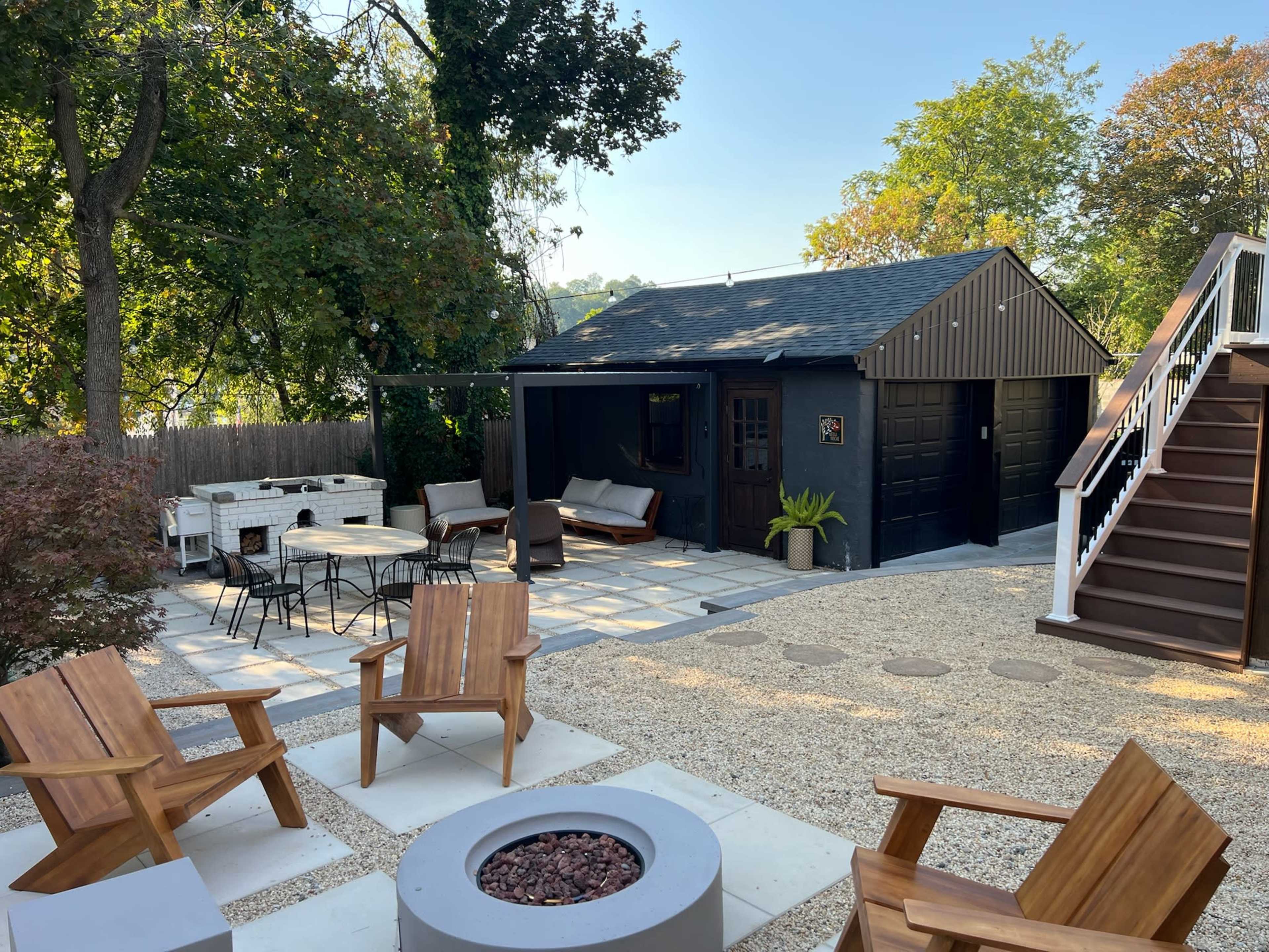 The backyard features a stone patio with a fire pit, surrounded by wooden chairs, and a garage-style building alongside a staircase leading to an upper deck area.