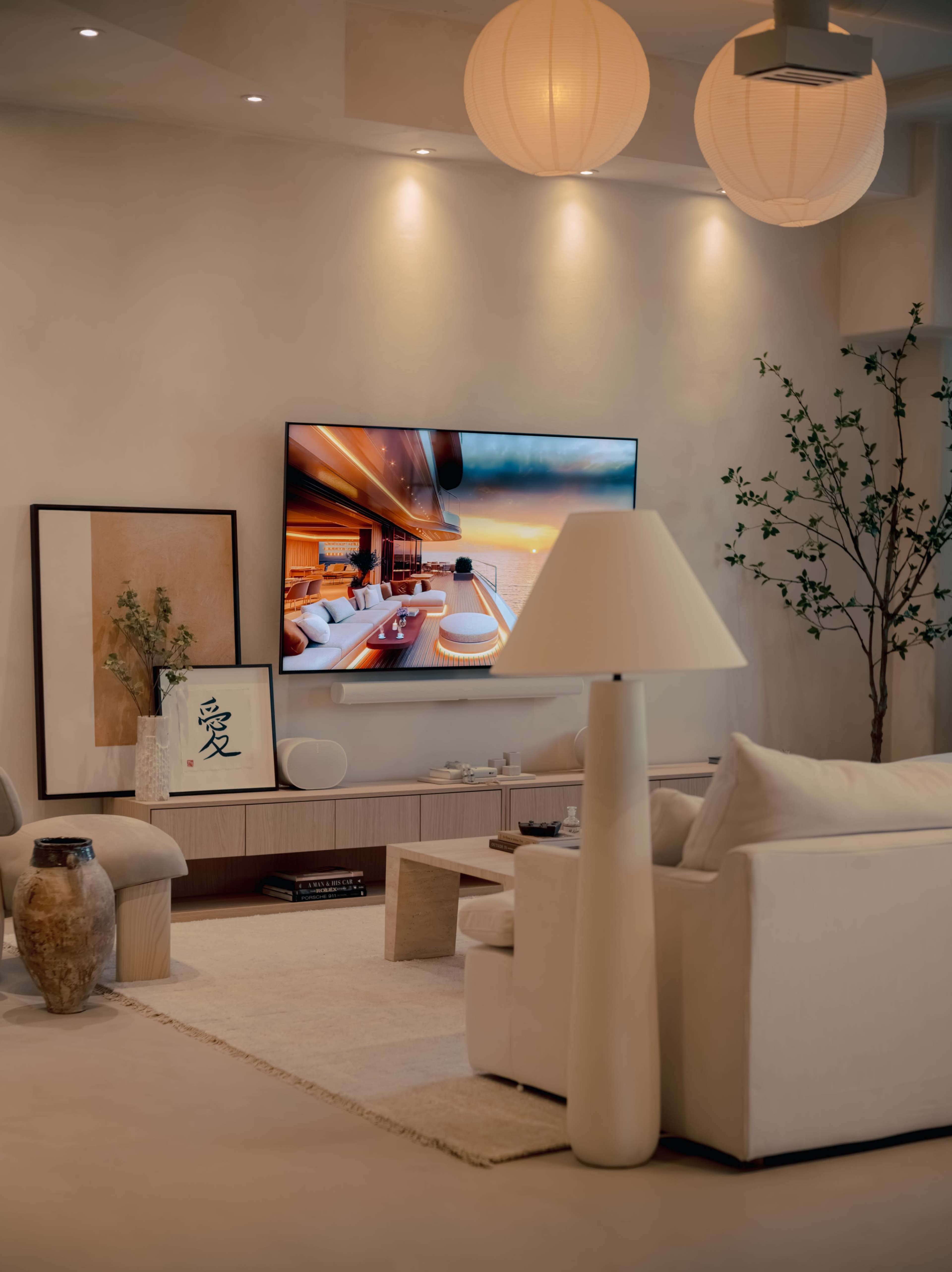 A modern living room features a light-colored sofa, a wooden coffee table with decorative elements, a large wall-mounted TV, and soft ambient lighting from ceiling fixtures.