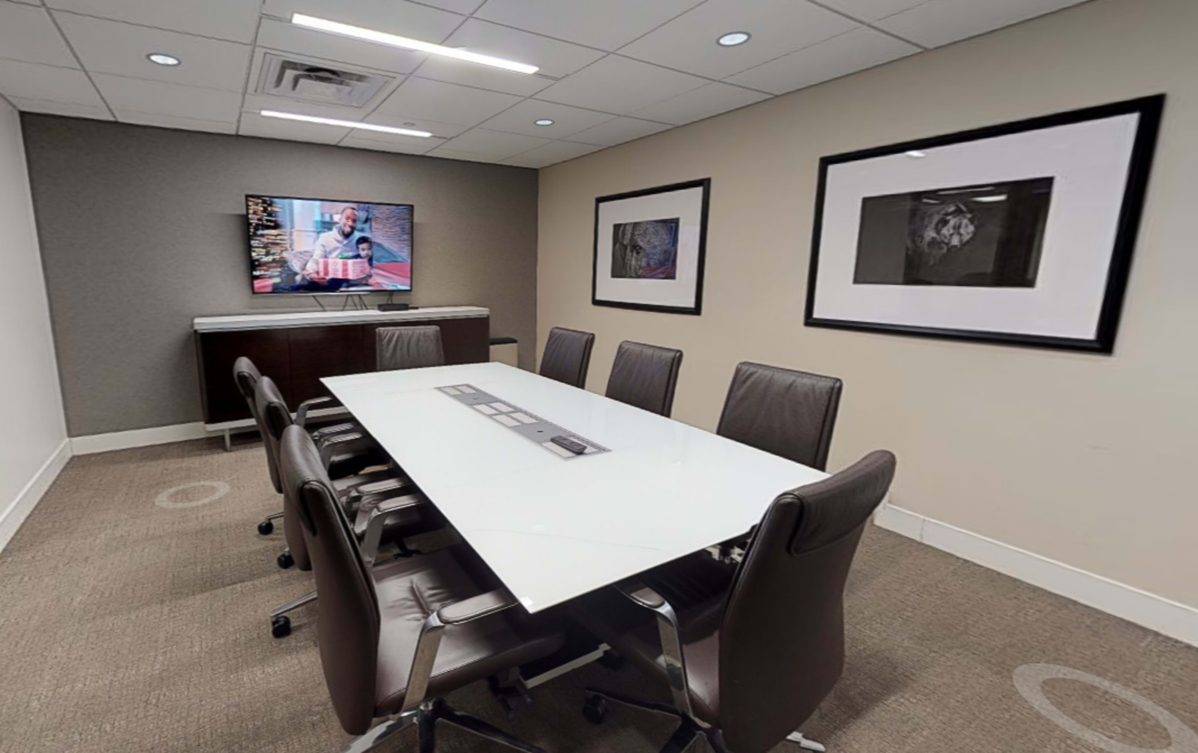 A modern conference room features a long glass table surrounded by eight leather chairs, with a wall-mounted television displaying content and framed artwork on the walls.
