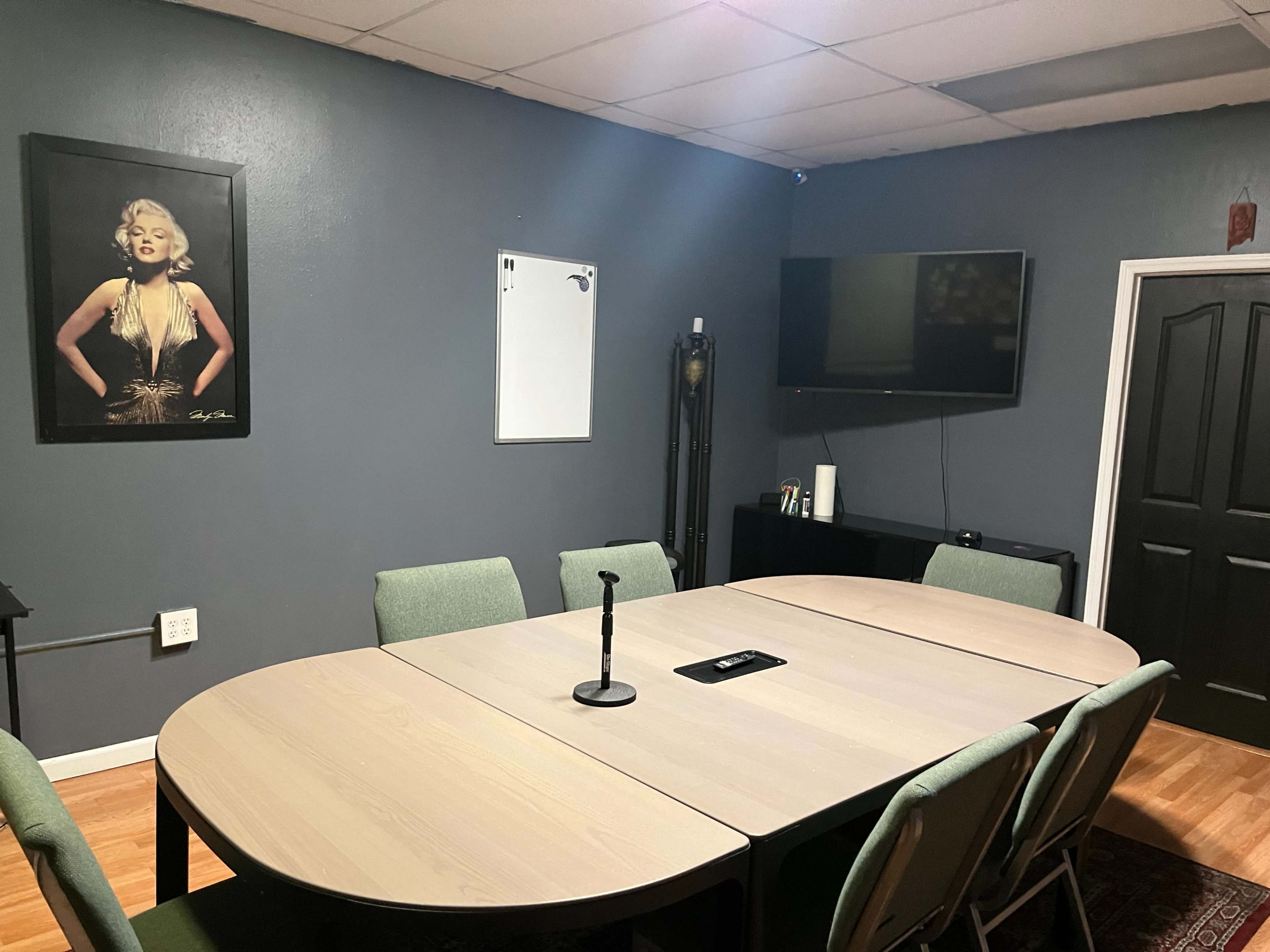 A conference room with a large table surrounded by chairs, a whiteboard on the wall, a television mounted on the opposite wall, and a framed portrait of a woman.