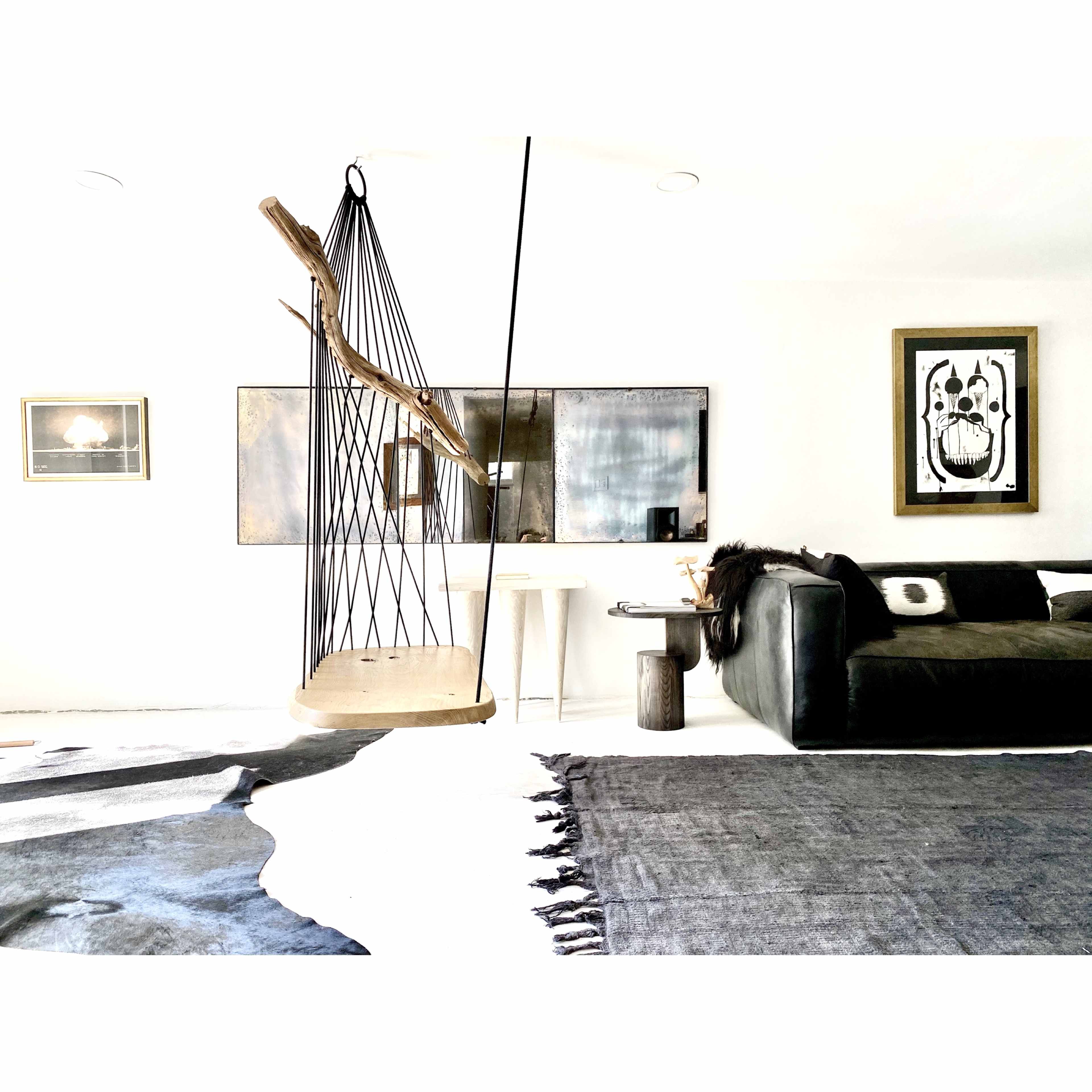 A modern living room featuring a swing chair suspended from the ceiling, a dark sofa, a small side table, and two framed artworks on white walls.